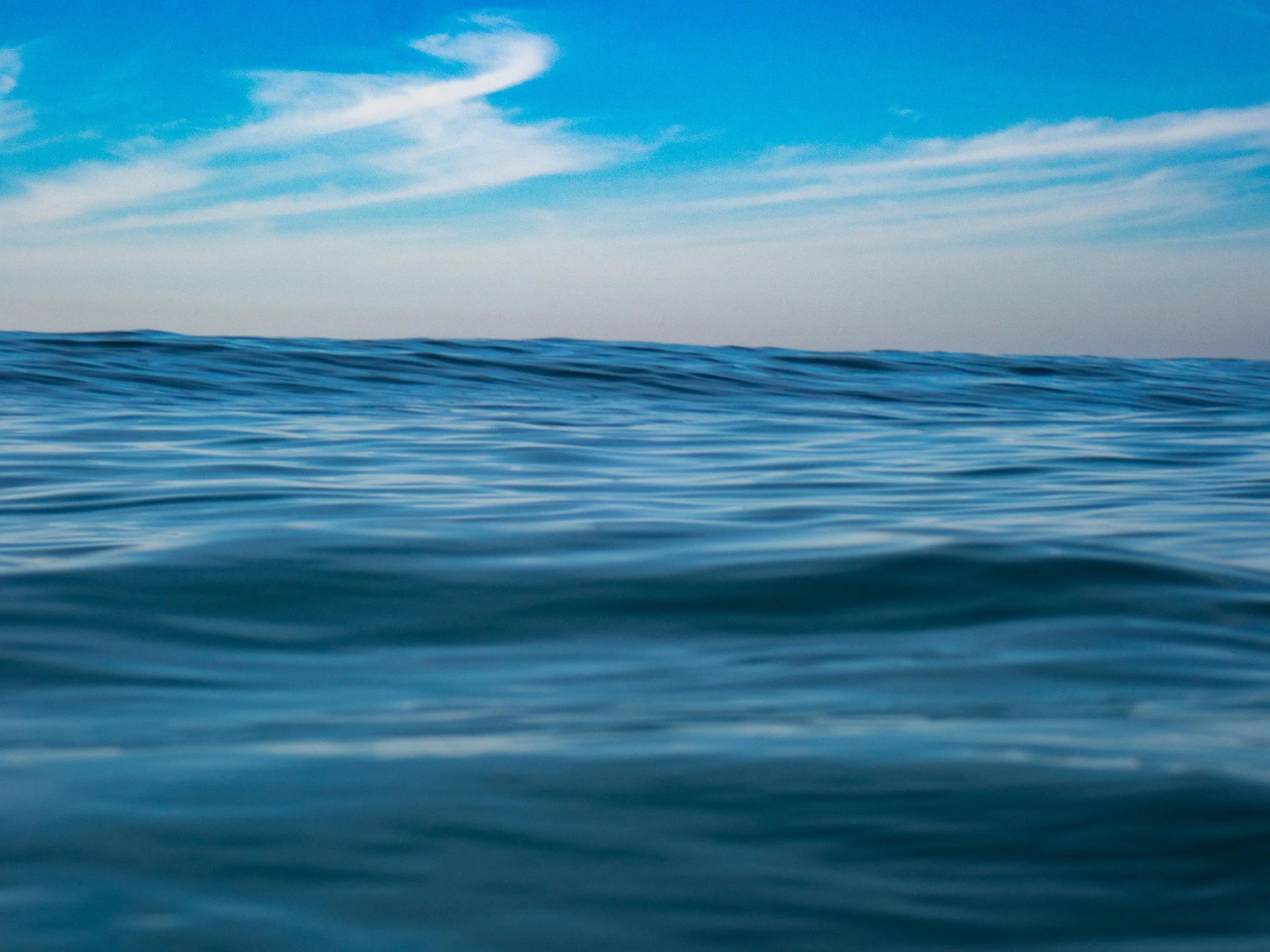A view of the open ocean with gentle waves under a bright blue sky with wispy clouds.
