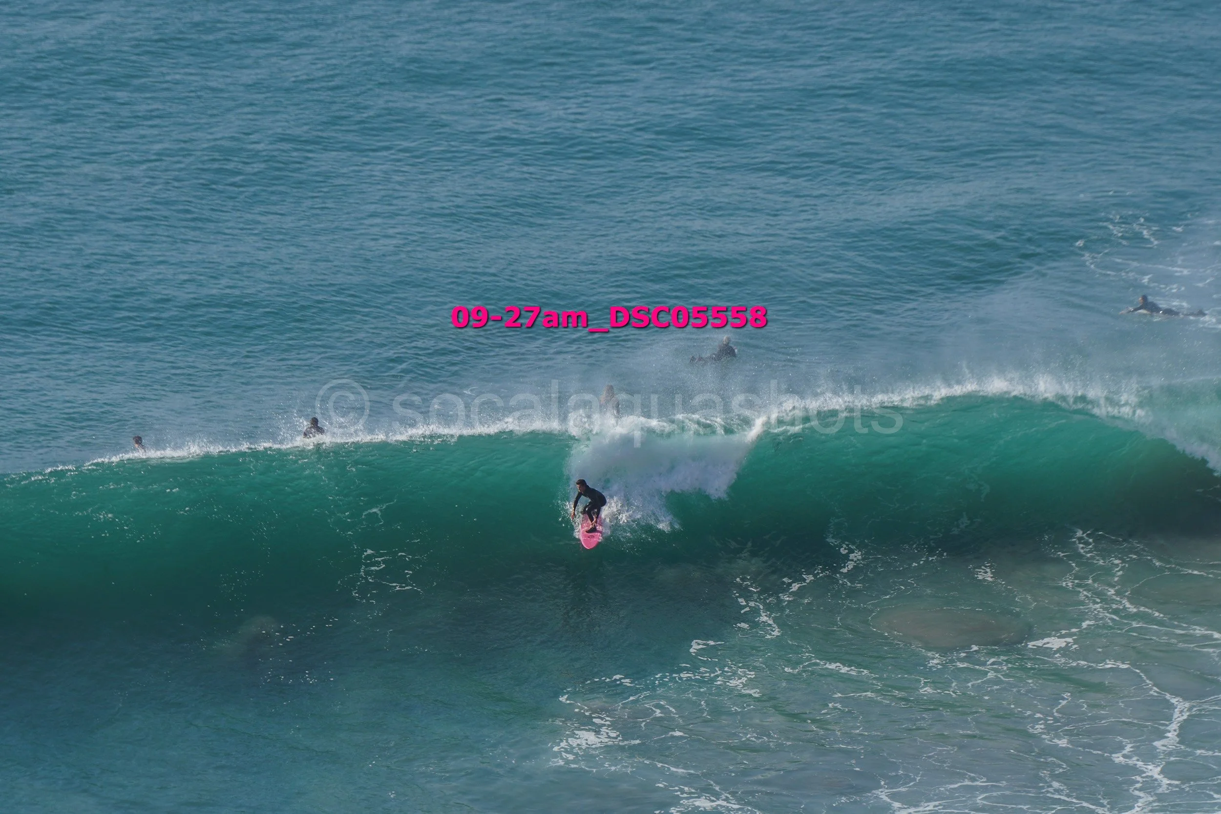 A person surfing on a pink surfboard riding a large wave with several other surfers in the water nearby.
