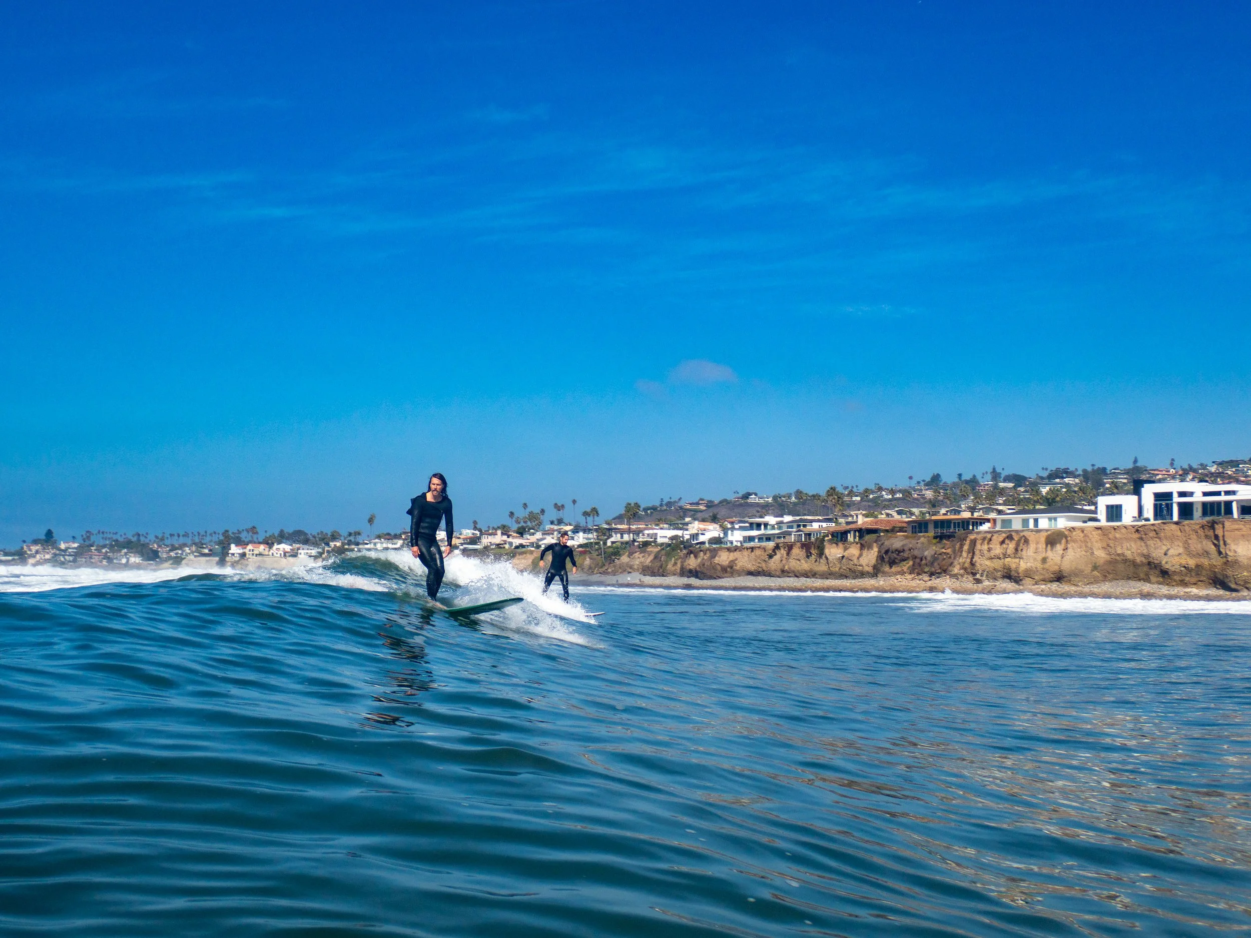 Two people surfing in the ocean with a coastal neighborhood and cliffs in the background on a clear day.