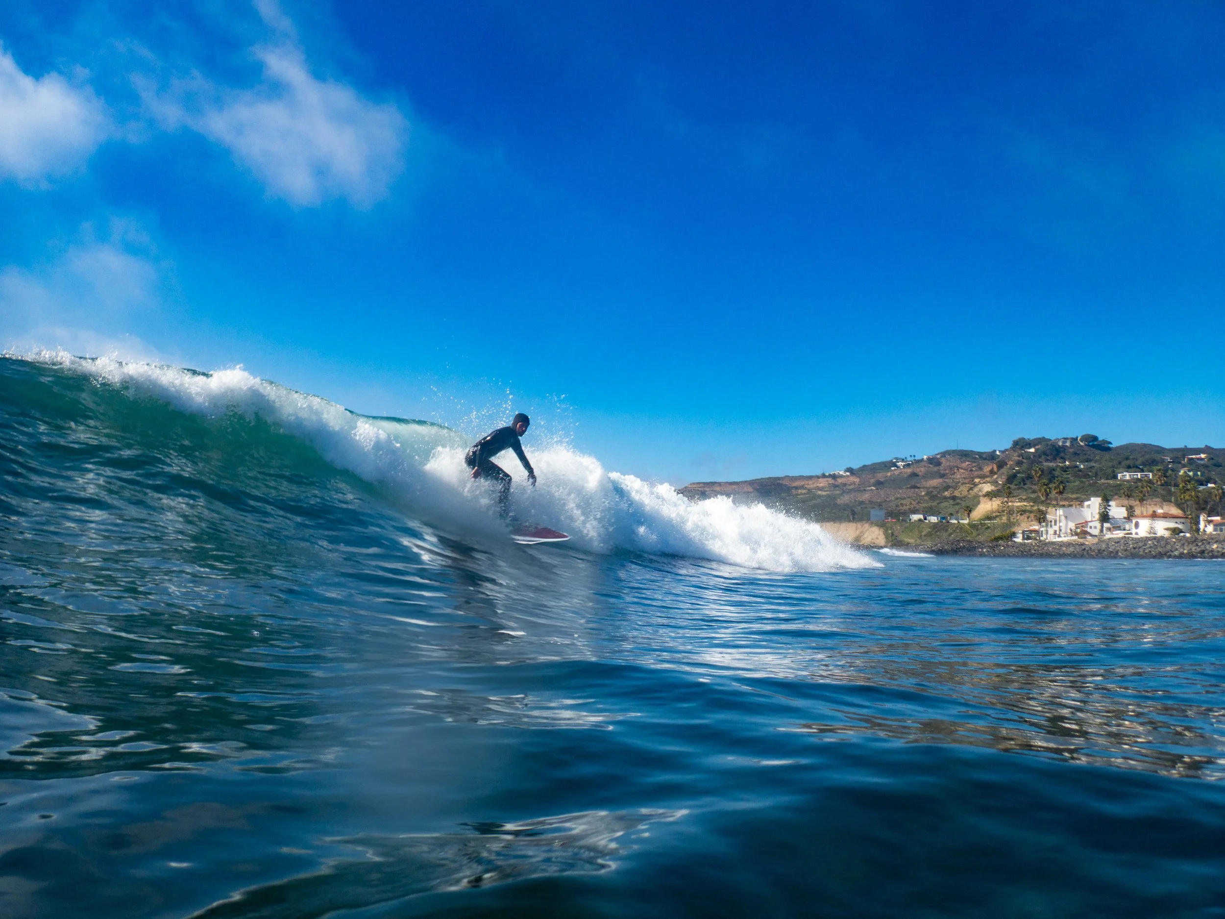 A person surfing on a wave in the ocean near a coastal town with houses and hills in the background under a clear blue sky.