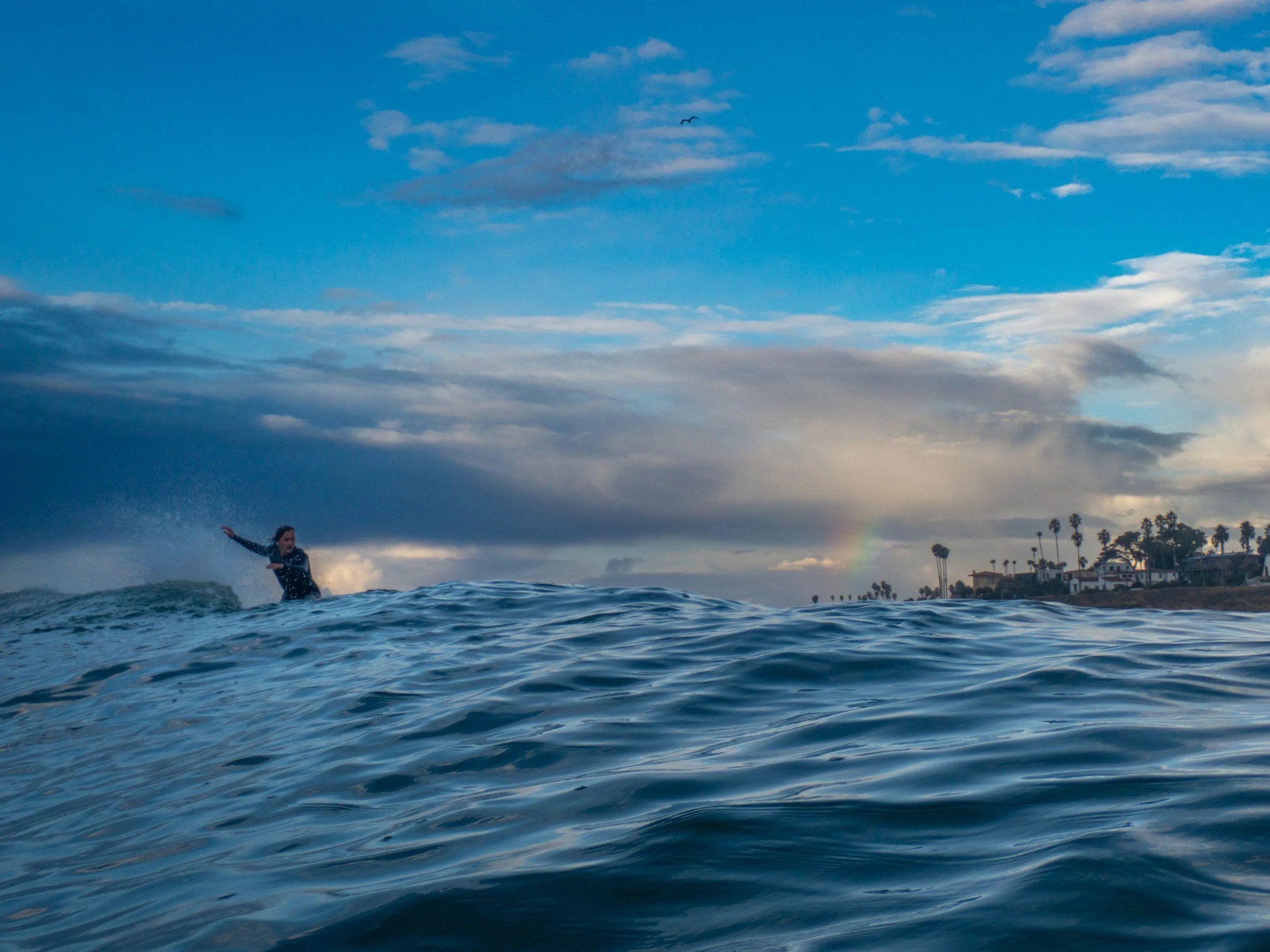 A person surfing on the ocean waves with a cloudy sky and a rainbow in the background.