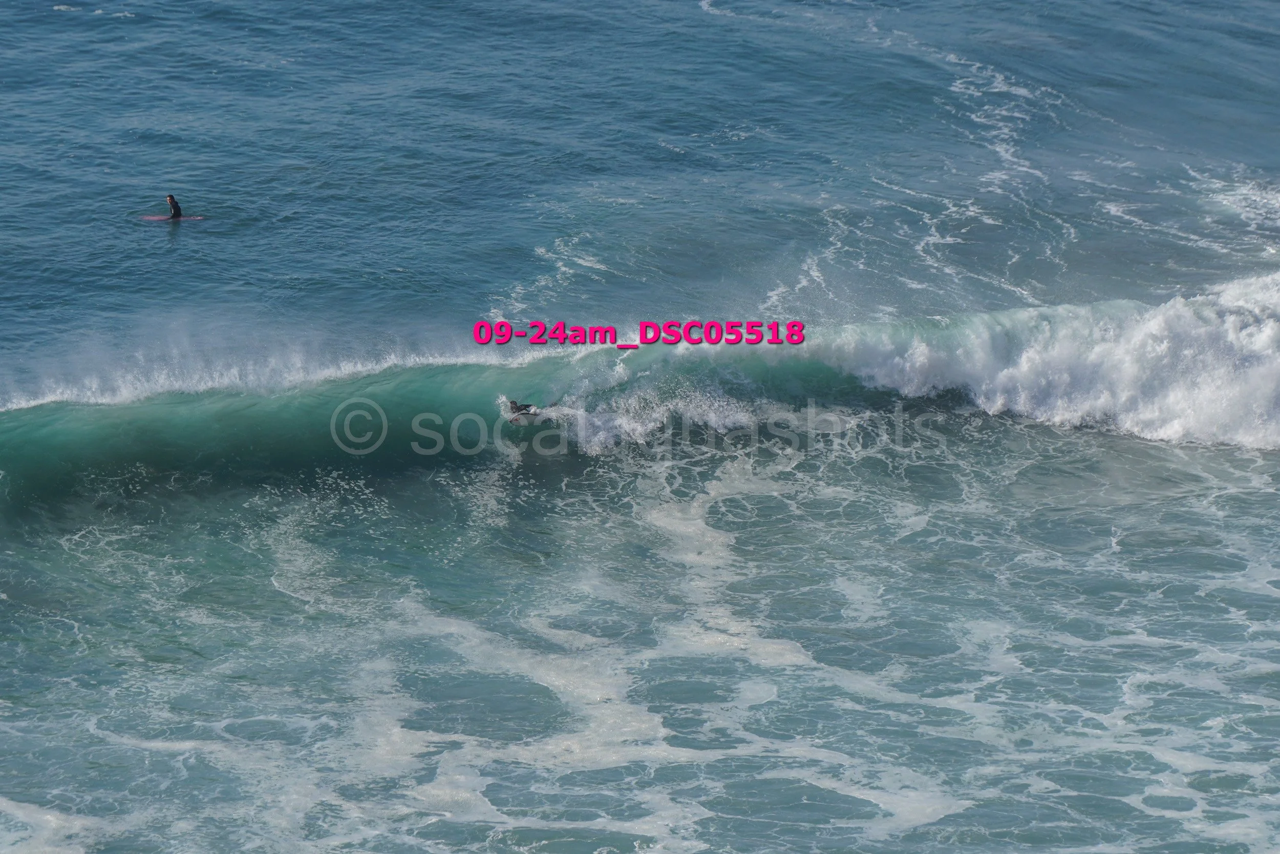 A person surfing on a wave in the ocean, with another person on a paddleboard in the distance.