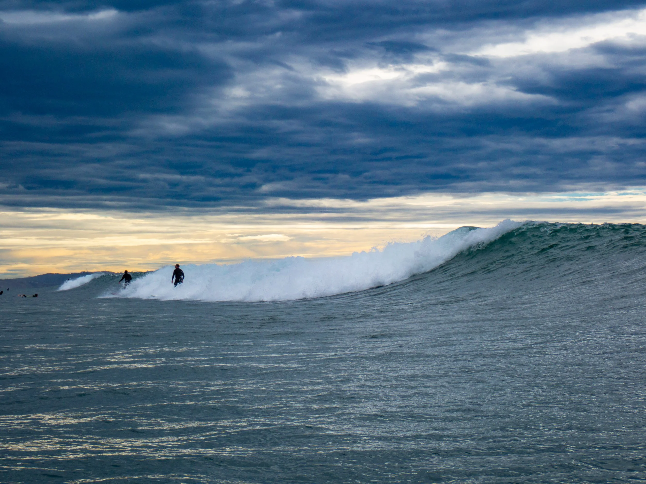Surfers riding waves in the ocean under a cloudy sky at sunset or sunrise.