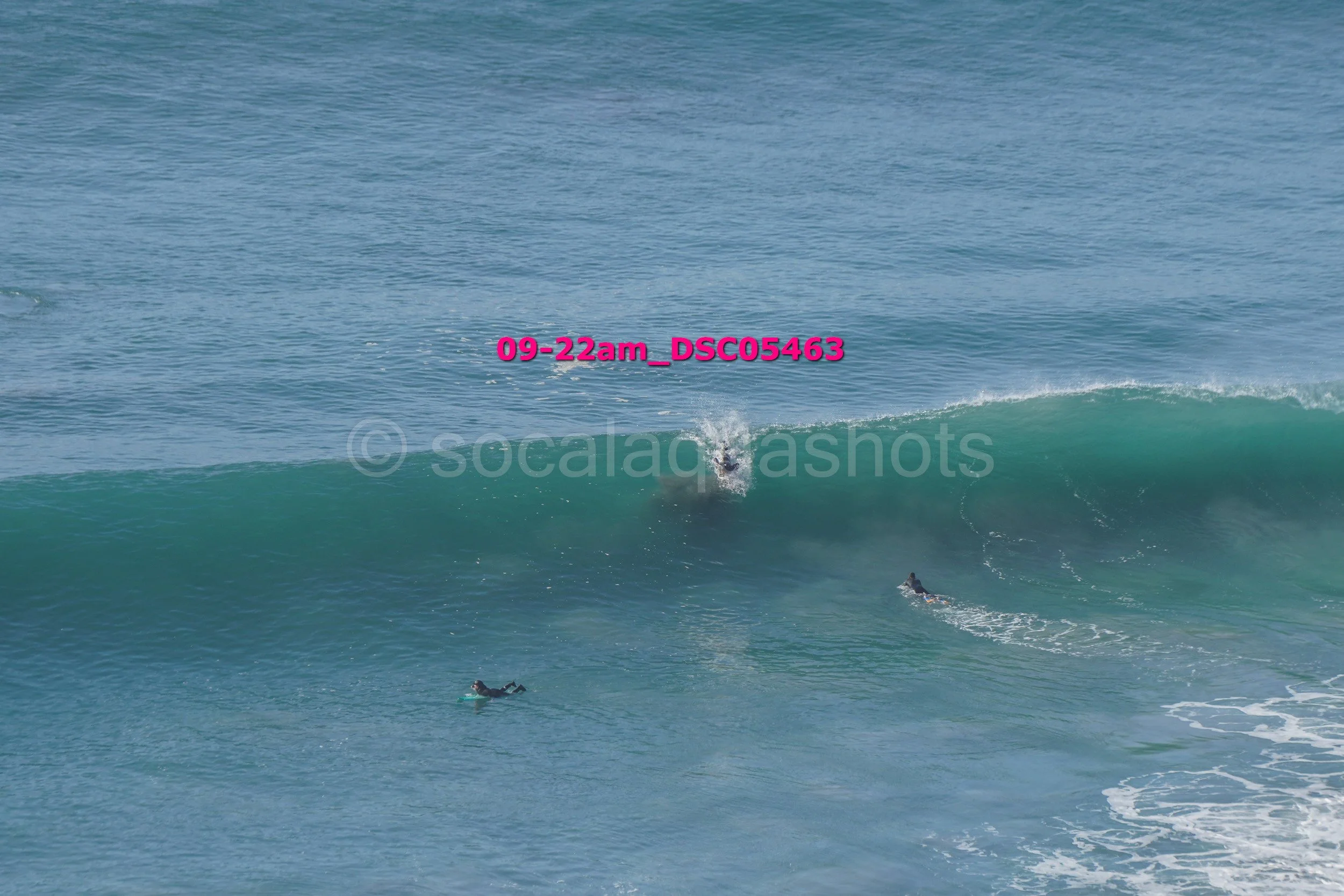 Two surfers in wetsuits riding ocean waves, one on the wave crest and another paddling in the water.