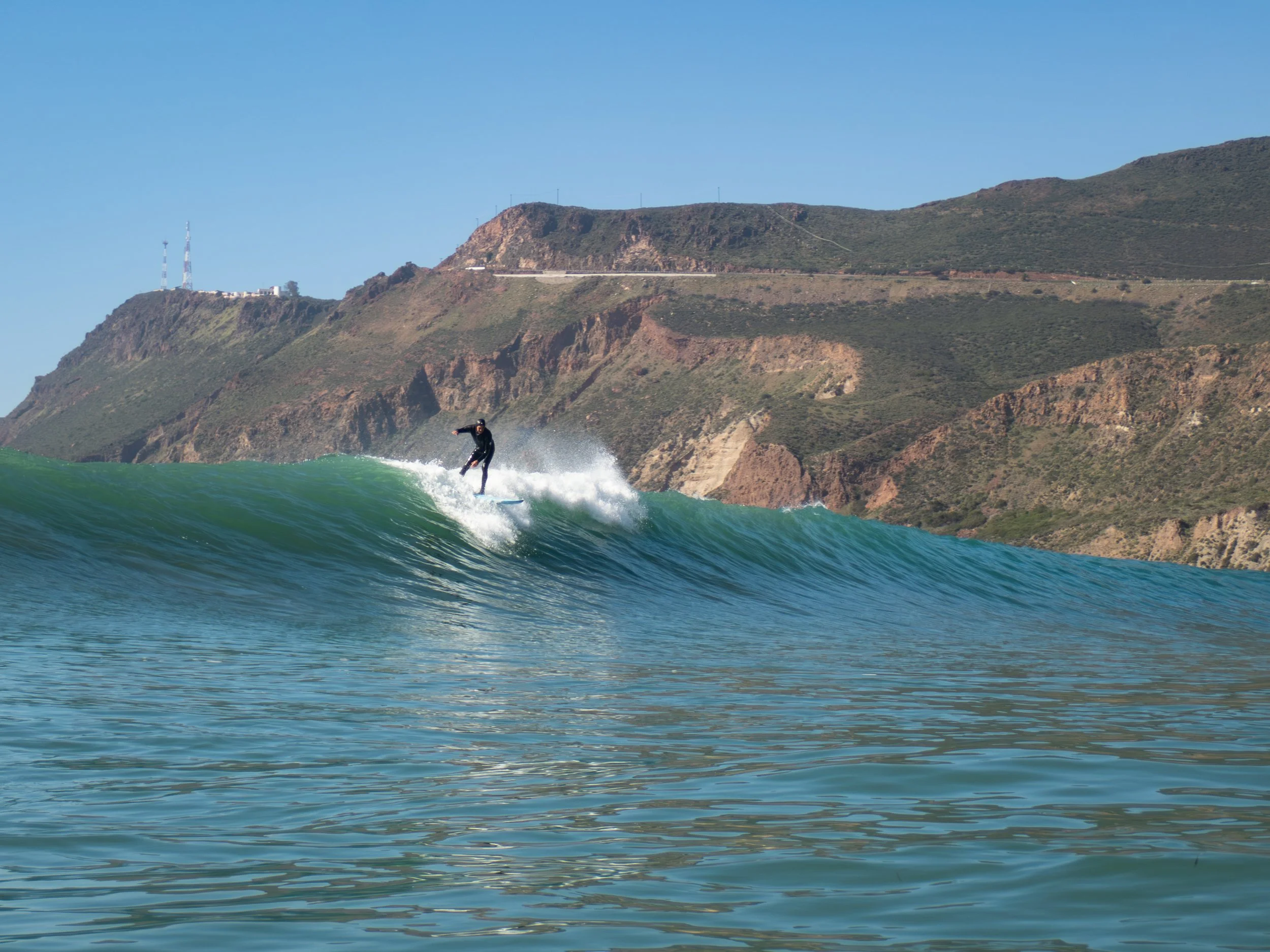 Surfer riding a wave near a mountainous coastline on a clear day.