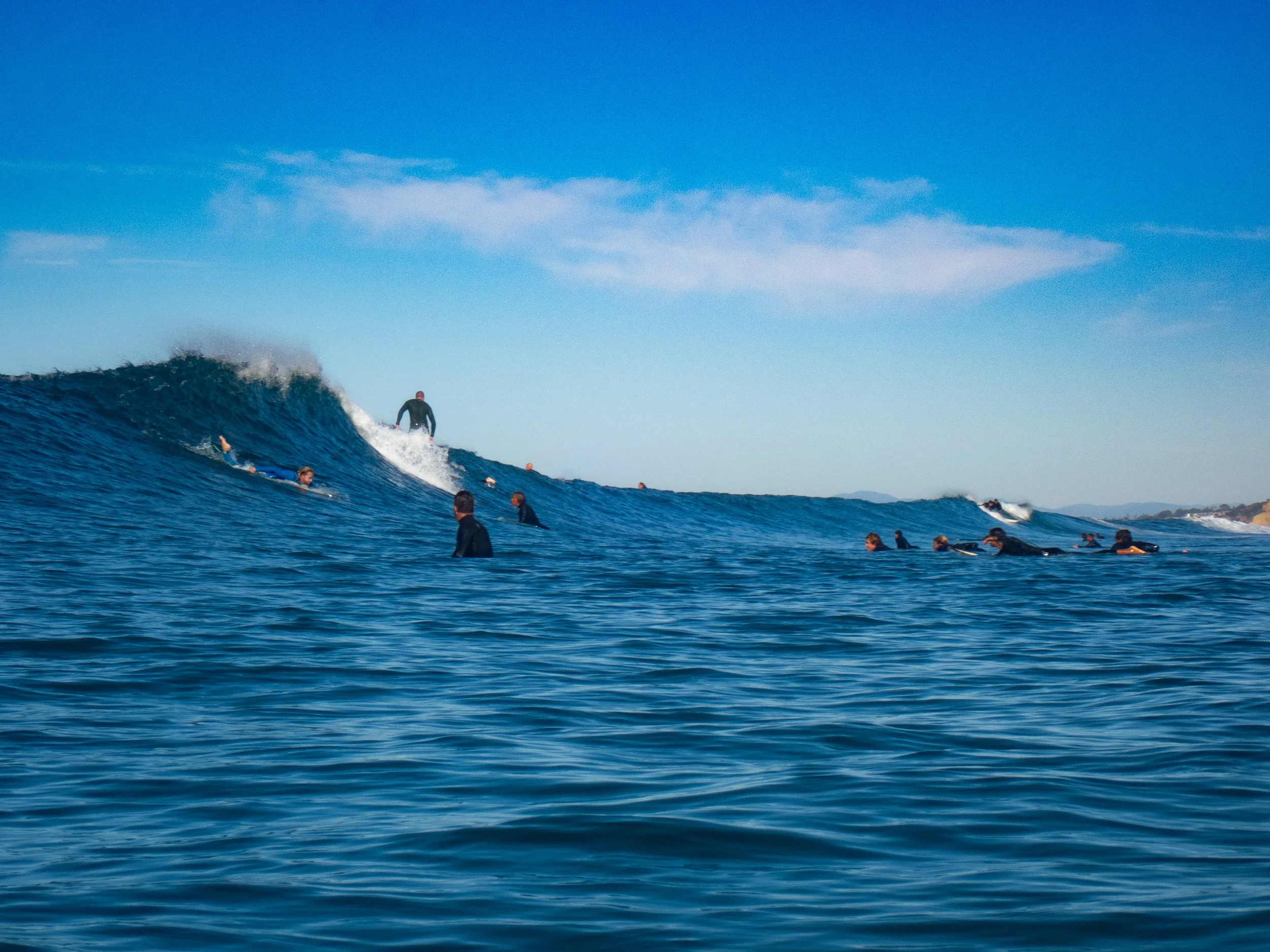 Surfers in wetsuits waiting in the water for waves at the beach, with one surfer riding a wave under a blue sky.