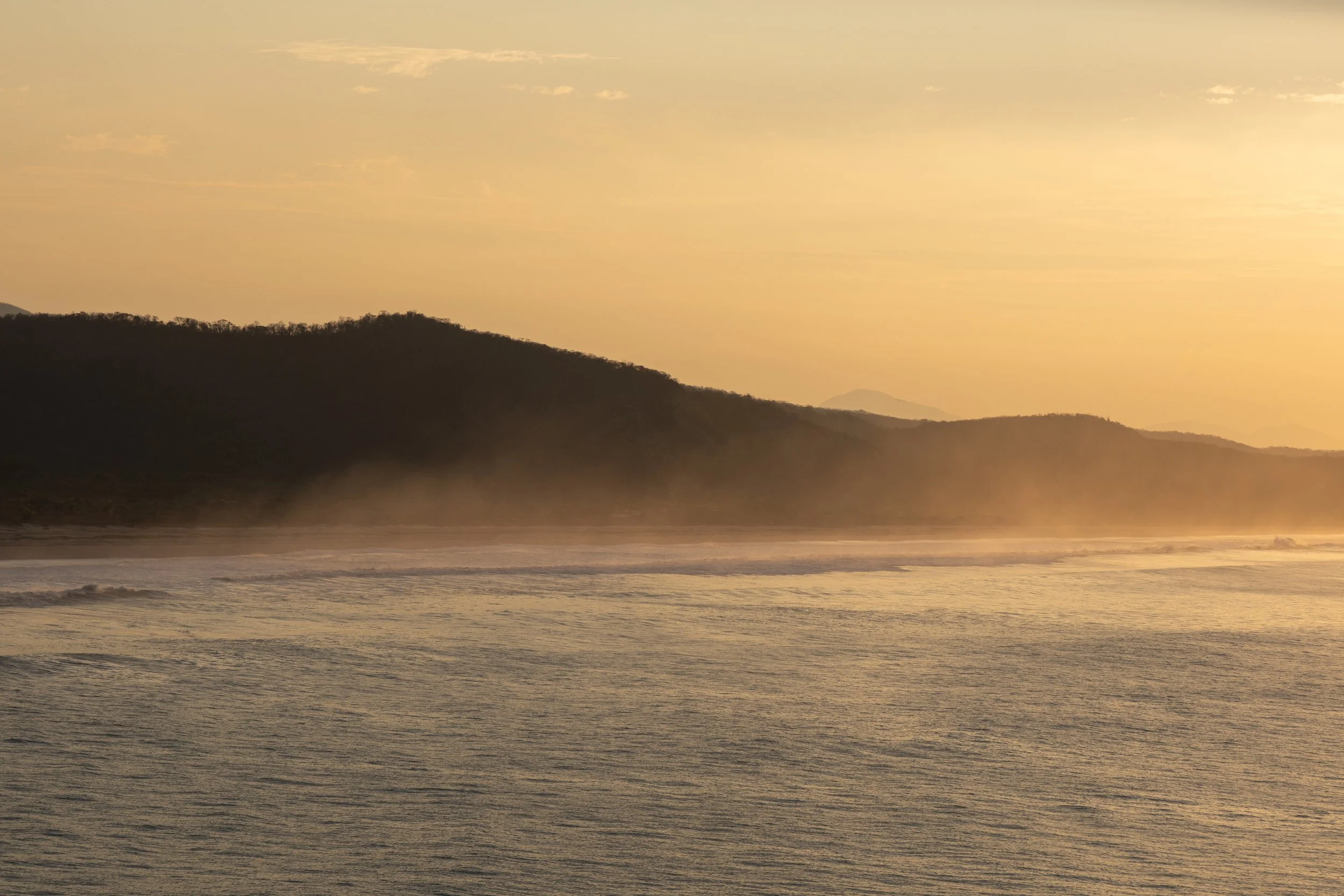 Sunset over a beach with mountain silhouettes in the background, calm waves in the foreground.