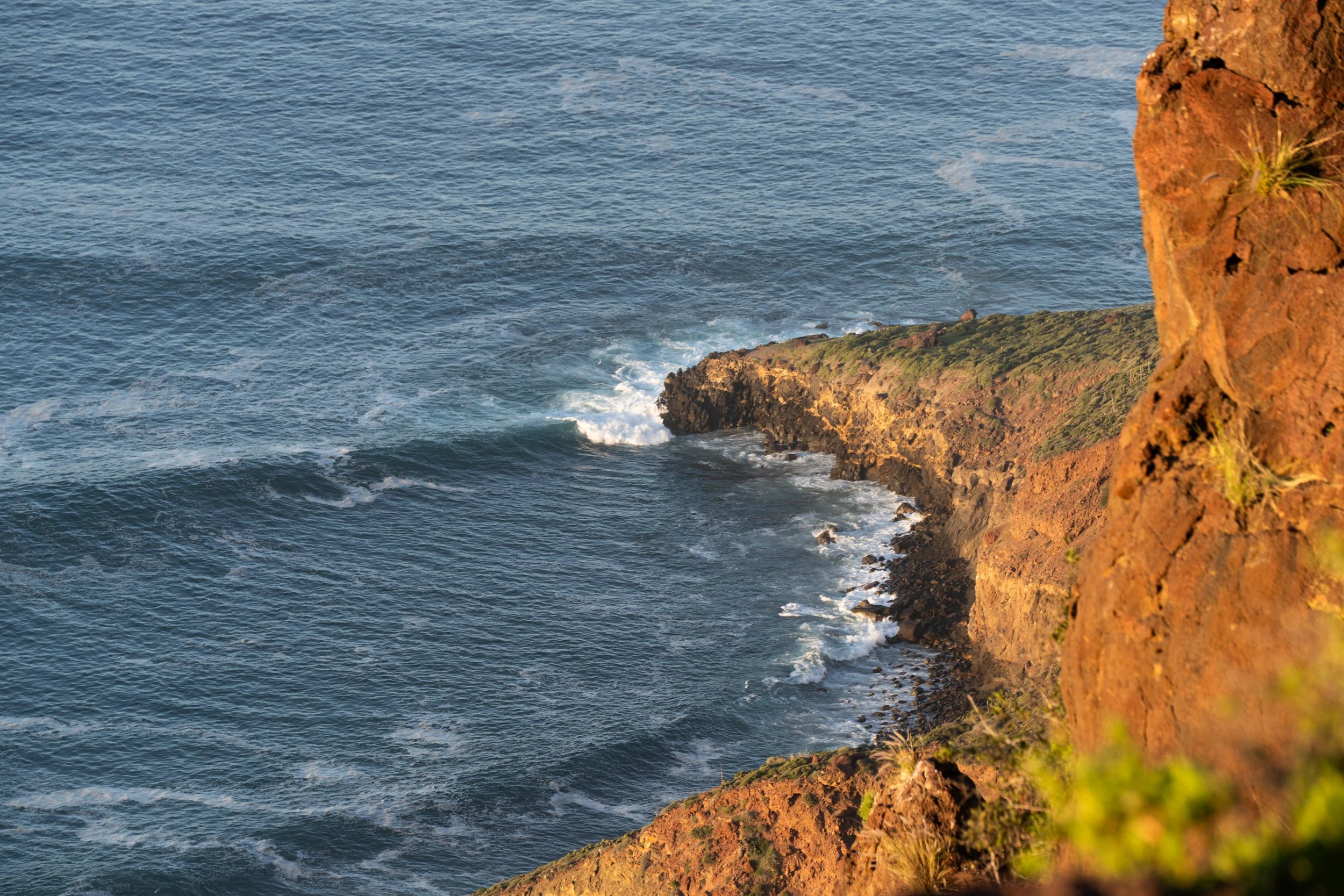 Cliffside view of the ocean with waves crashing against the rocky shore and a reddish-brown cliff in the foreground.