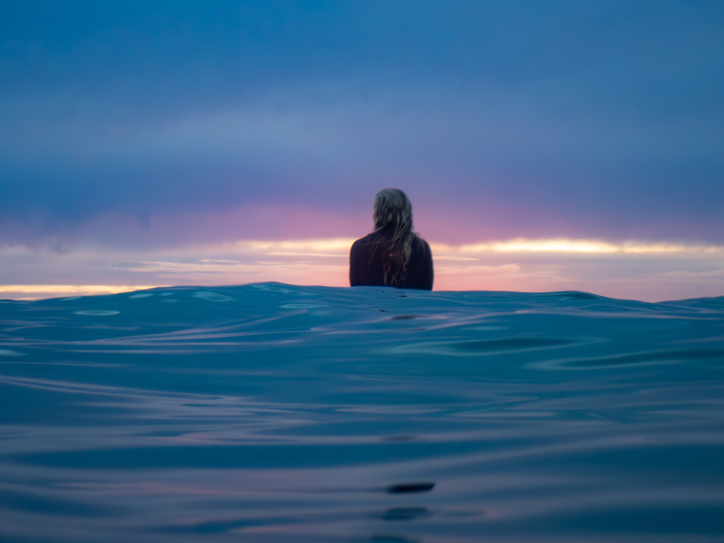 A person with long hair sitting in the ocean, facing a colorful sunset sky.