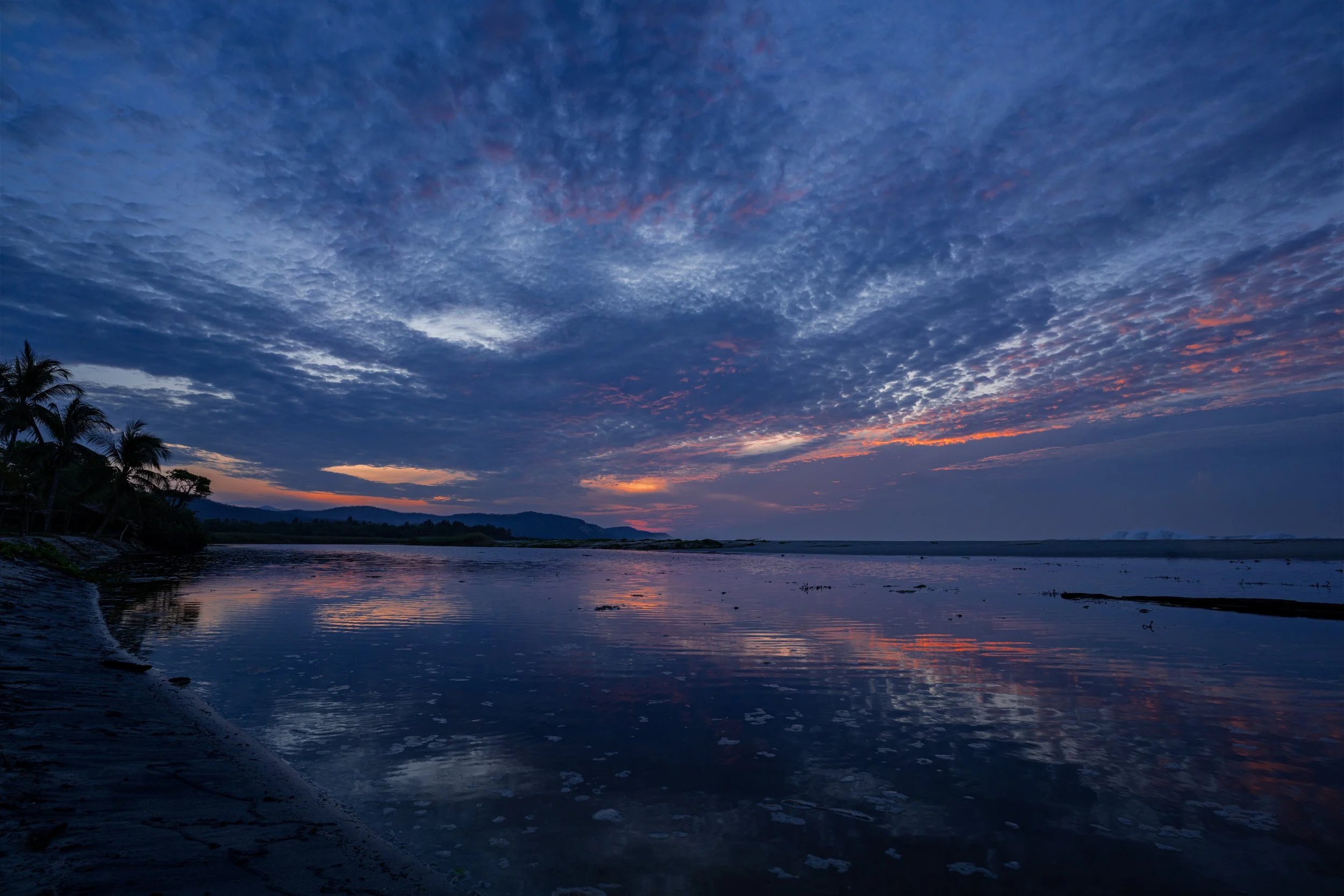 Sunset over a river with clouds in the sky and palm trees on the left side.