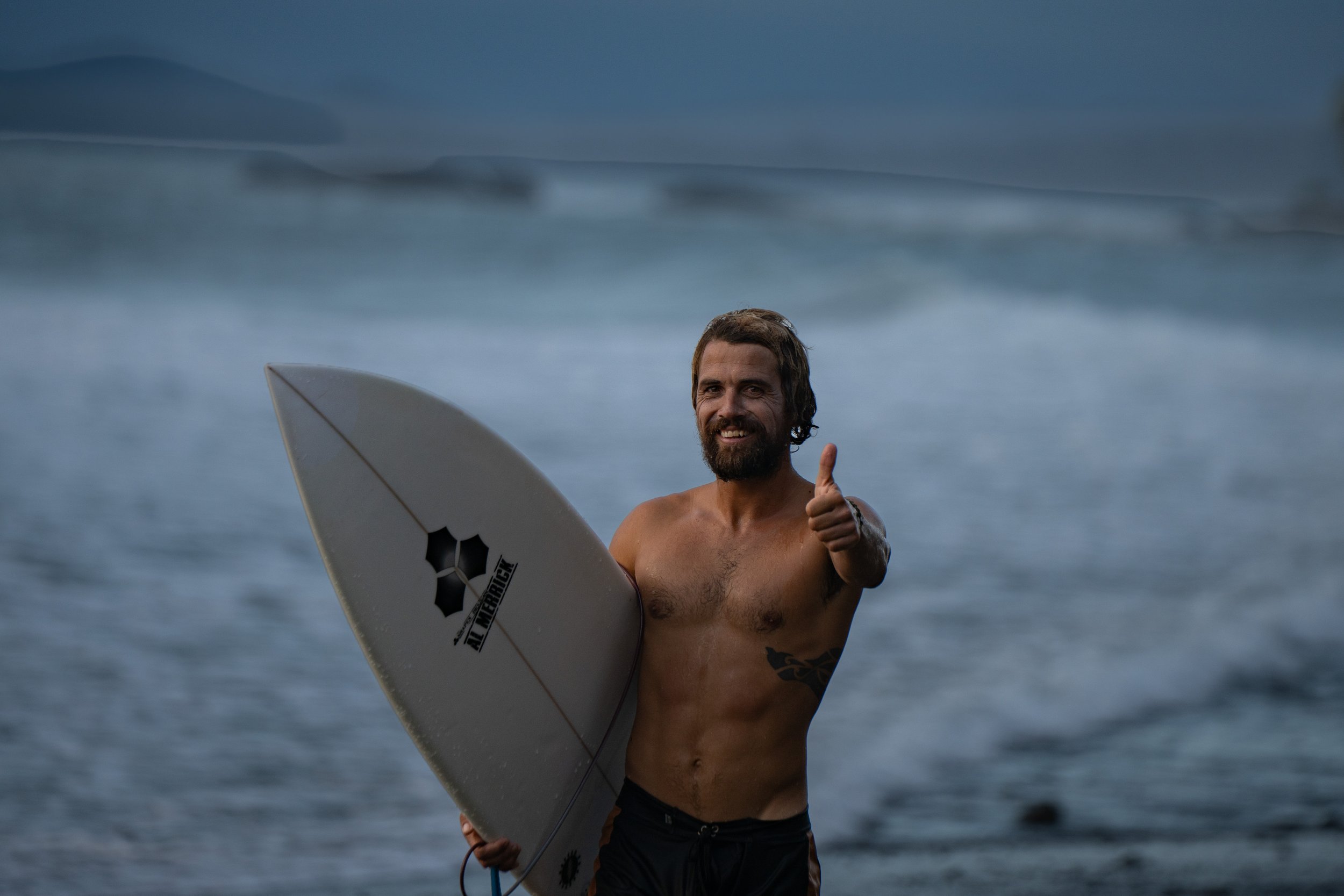 A shirtless man with a beard and long hair holding a surfboard and giving a thumbs-up on the beach with ocean and distant hills in the background.