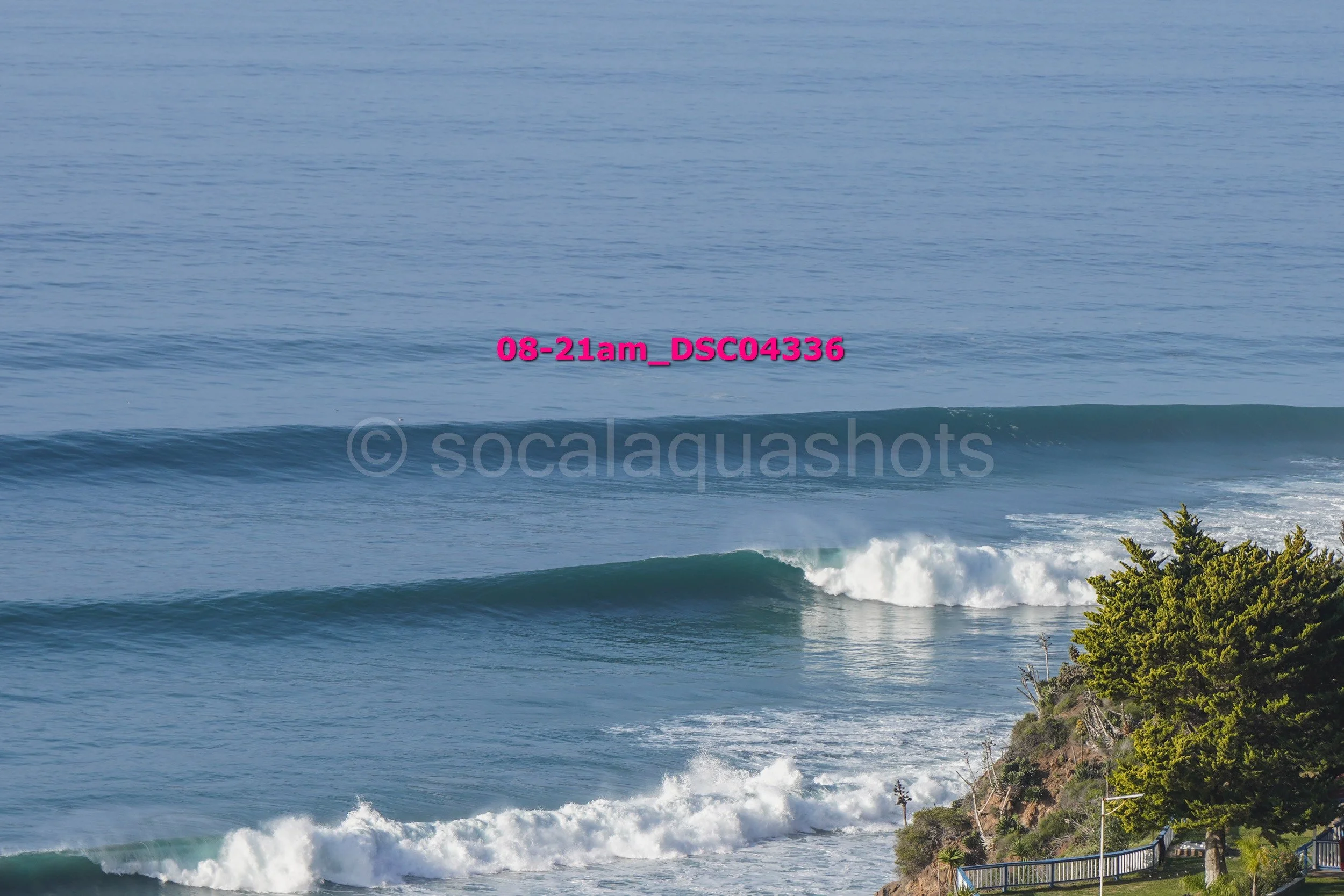 Ocean waves crashing near a rocky shoreline with greenery and trees.