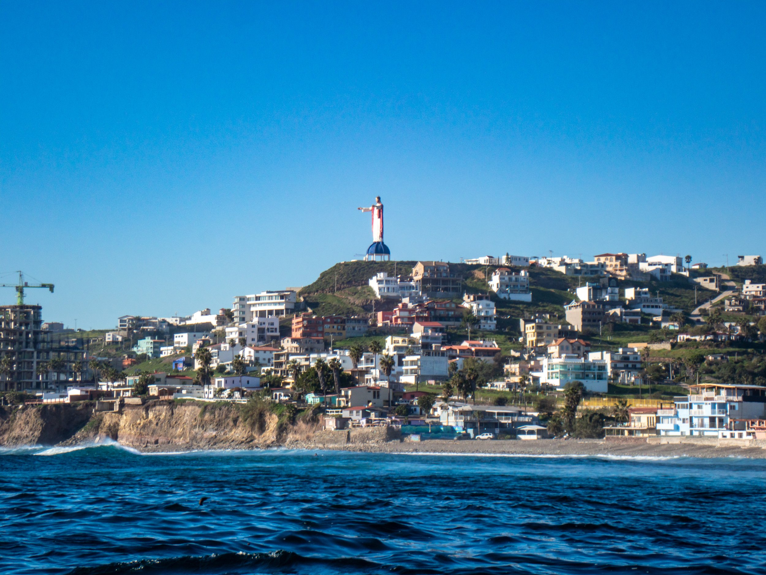 View of the San Diego coastline with the iconic Jesus statue on a hilltop and residential buildings below, under a clear blue sky.