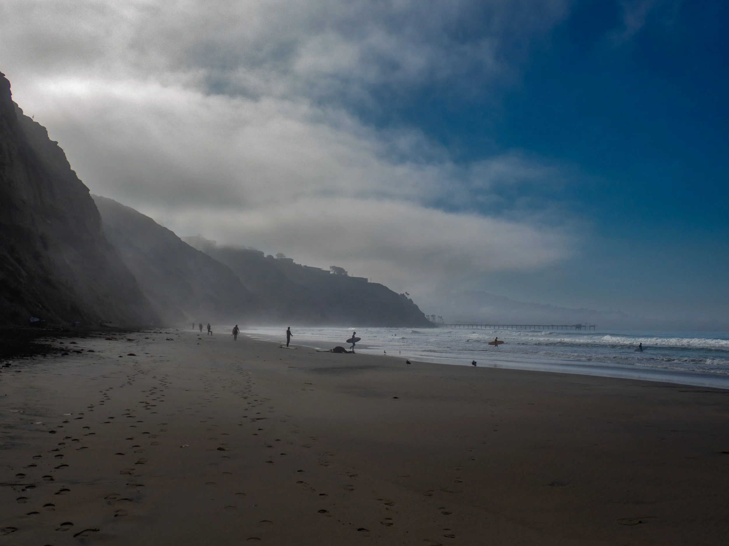 A sandy beach with a few people, some walking and others with surfboards, along a coastline with cliffs and a pier in the distance under a partly cloudy sky.