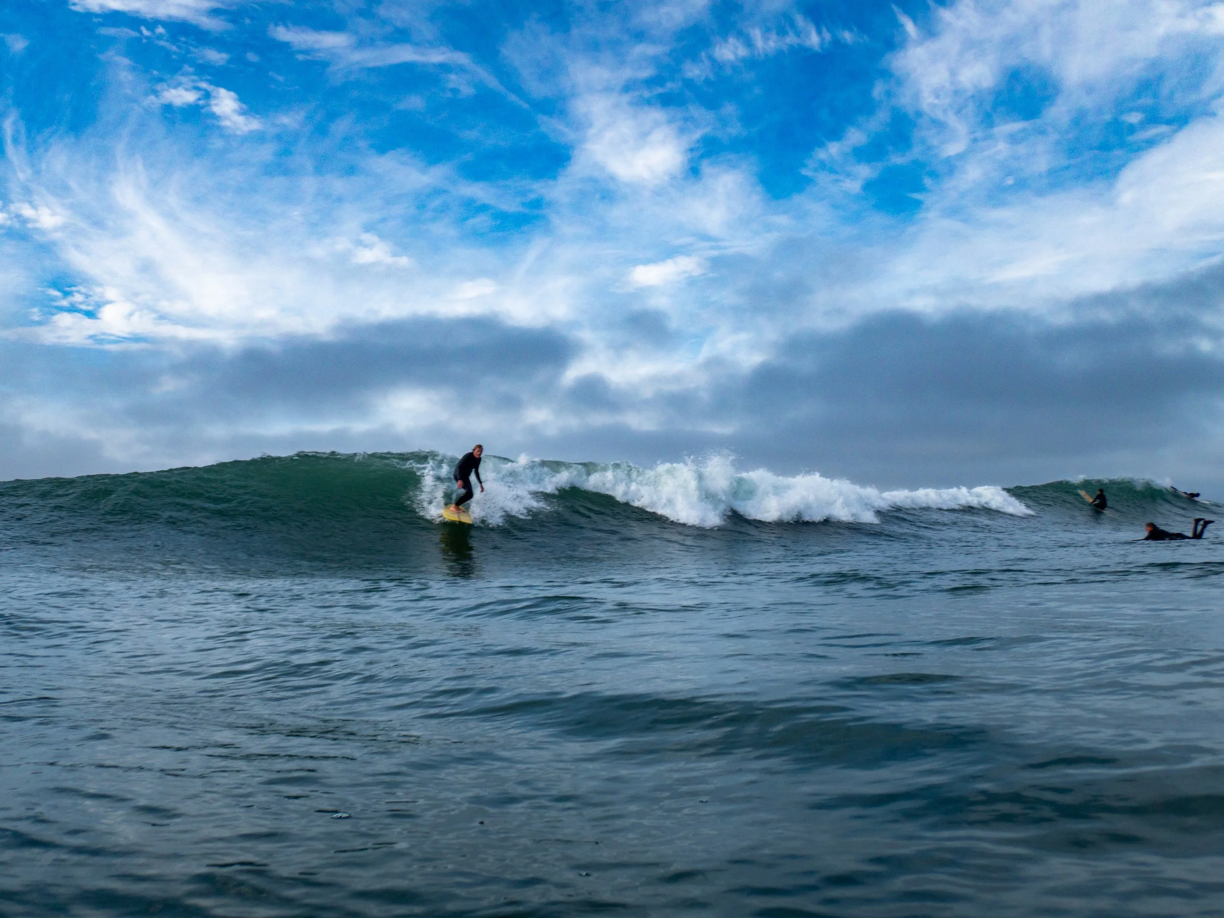 A person surfing on a wave in the ocean with a cloudy sky above and other surfers in the distance.