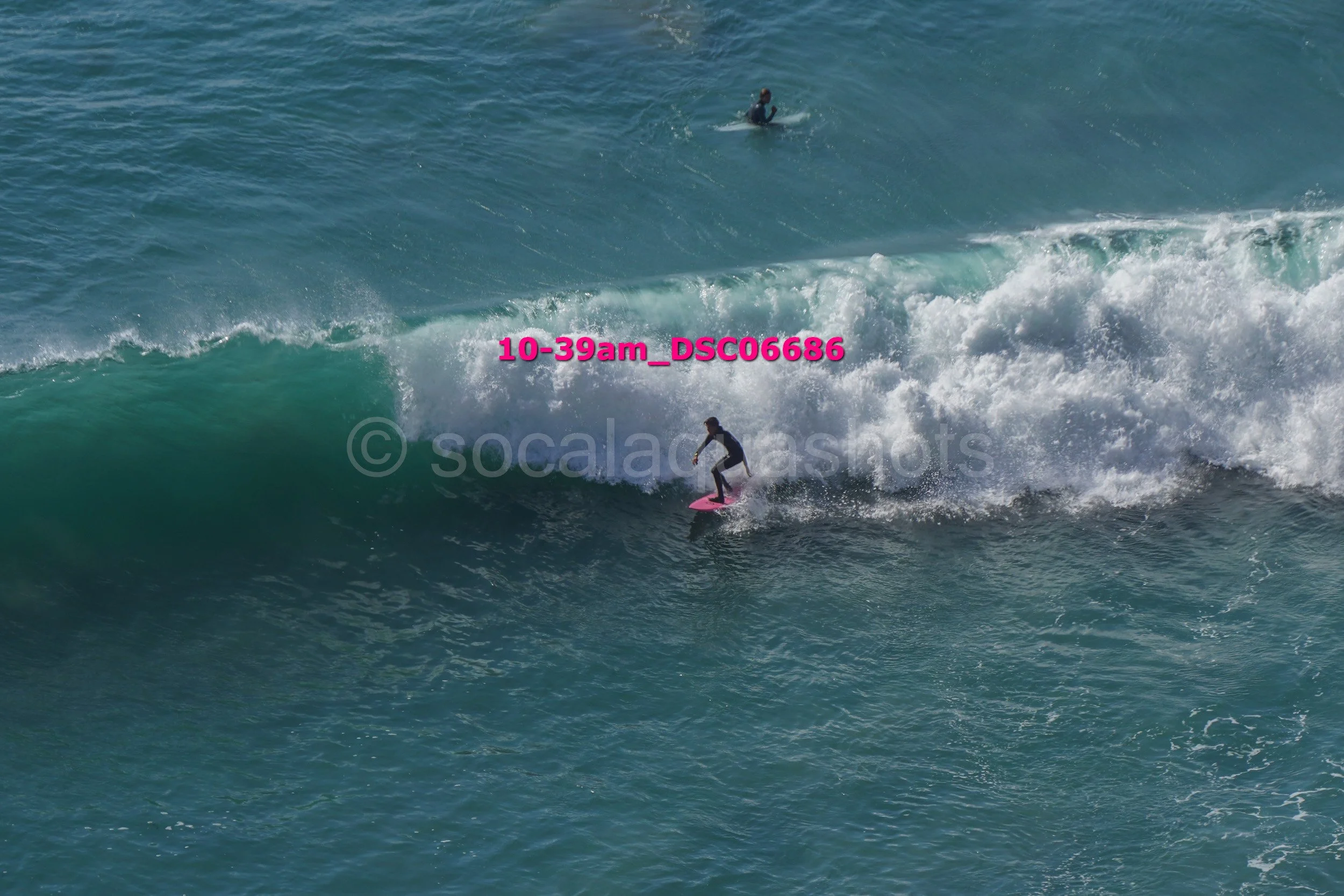 Person surfing on a pink surfboard on a large wave, another surfer is visible in the background in the ocean.