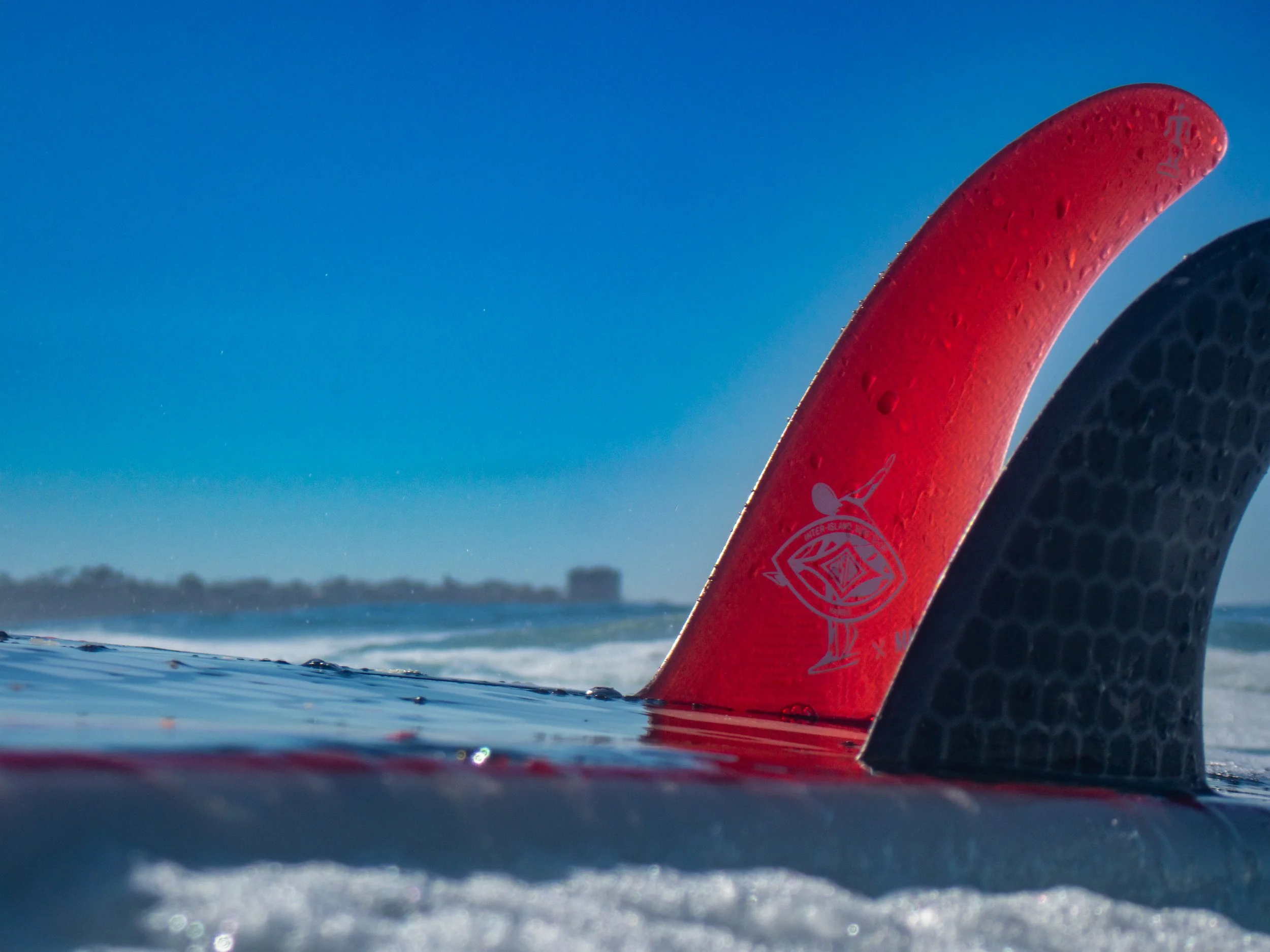 Close-up of a red and black surfboard fins sticking out of the water on a beach, with a clear blue sky and ocean waves in the background.