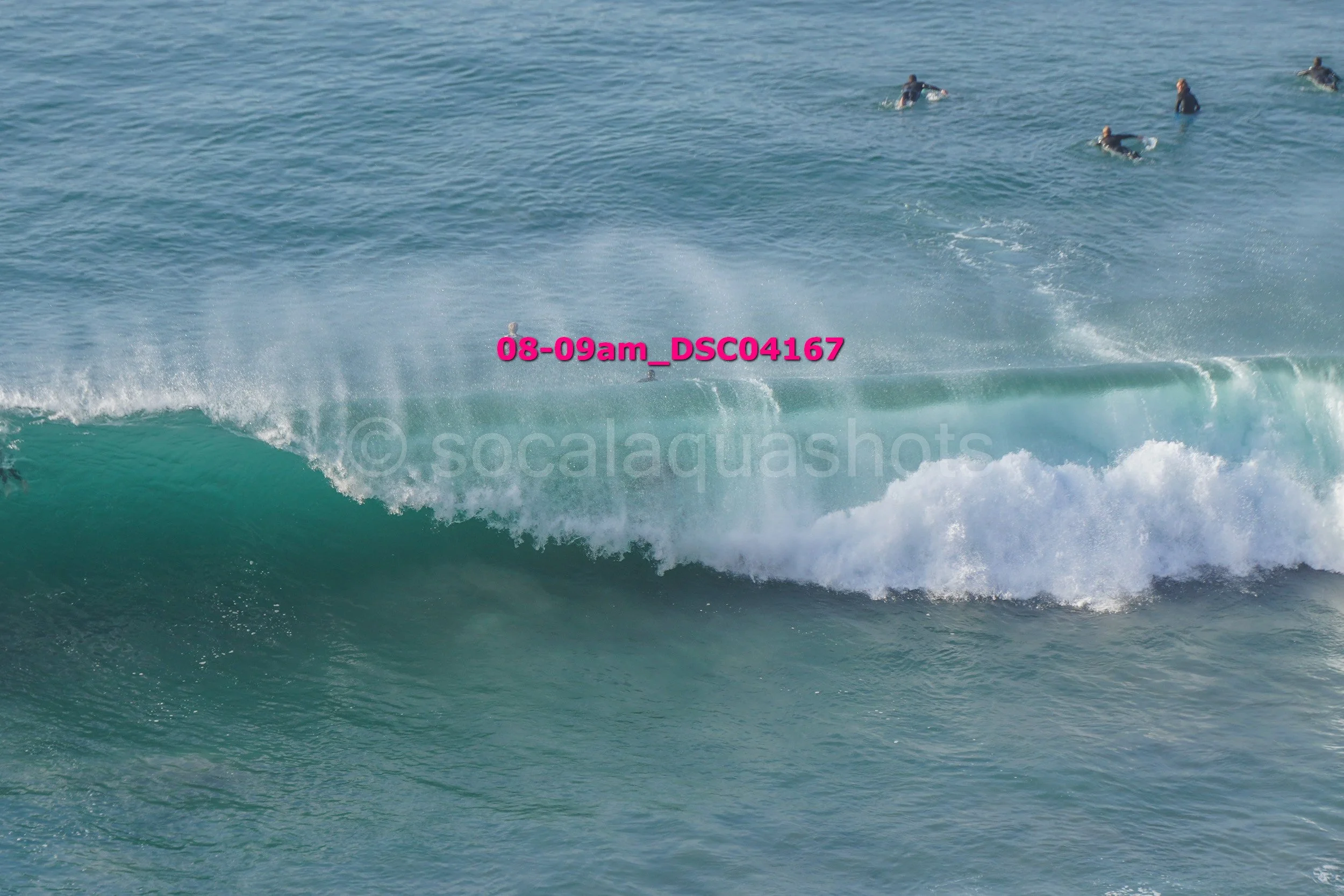 A surfer riding a large wave in the ocean with several people swimming in the background.