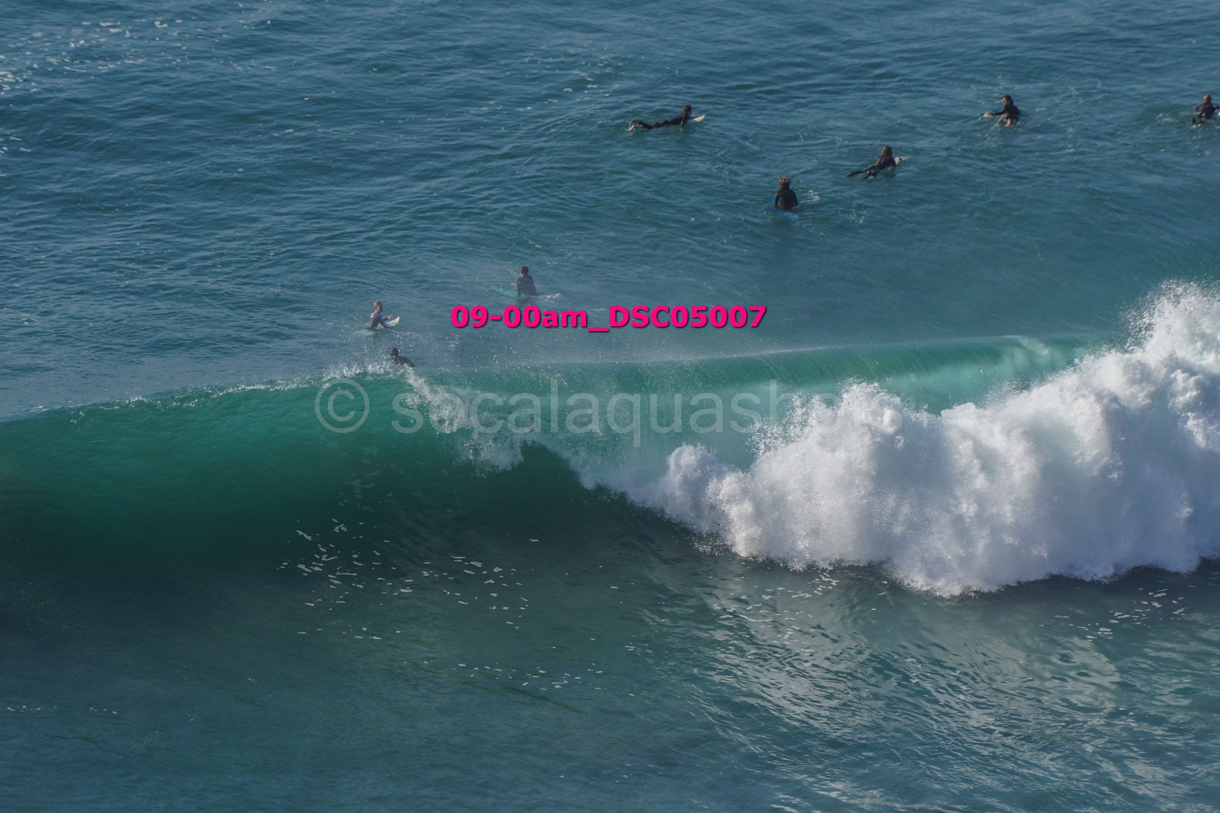 People in wetsuits surfing in the ocean, with one surfer on a wave and others swimming or floating nearby.