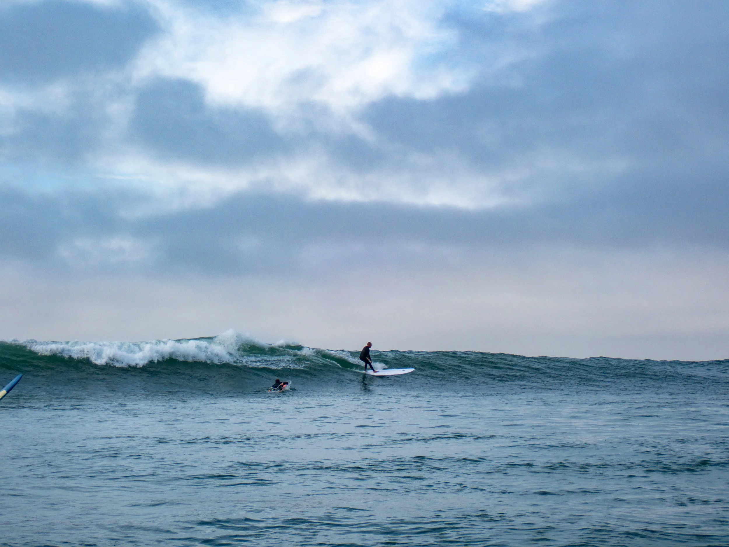 A person surfing on a wave in an ocean under a cloudy sky with two other surfers floating in the water.