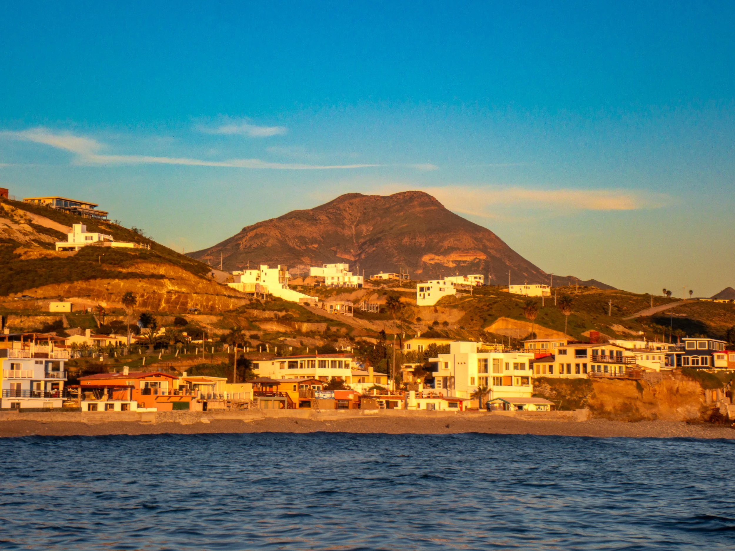Coastal view of hillside residential houses at sunset with a mountain in the background and water in the foreground.