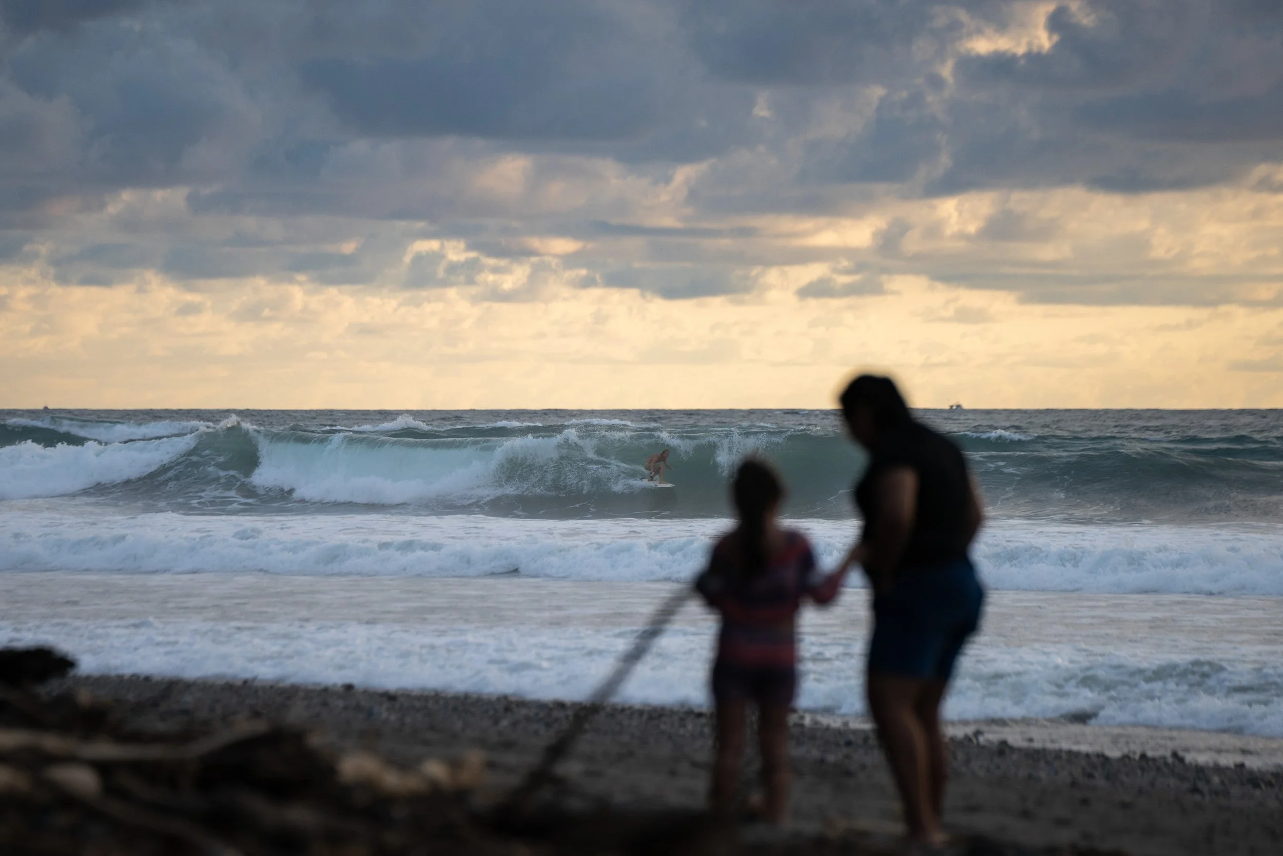 People on the beach near the ocean, with an individual surfing a wave in the distance, under a cloudy sky at sunset.