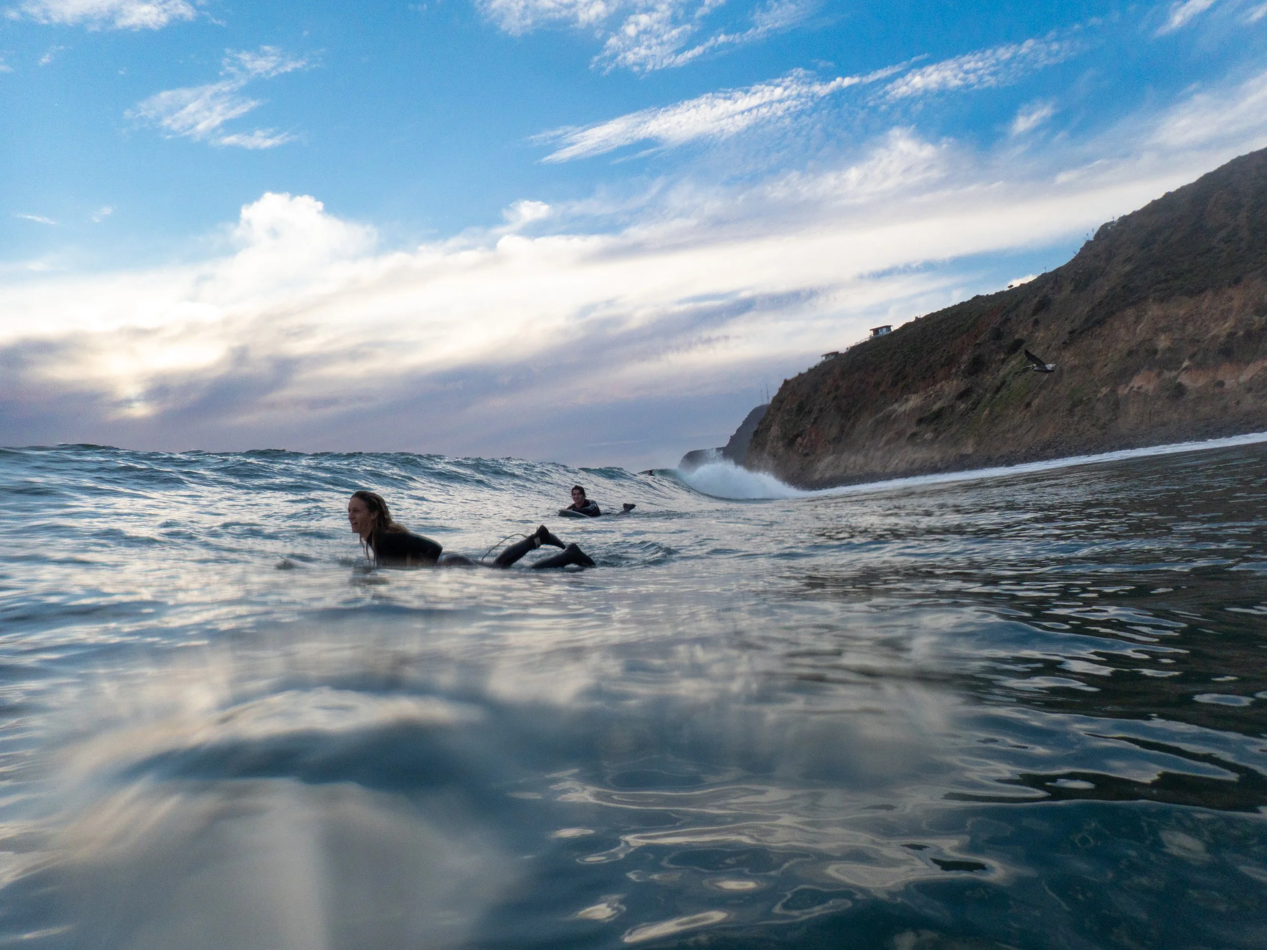 People surfing in the ocean near a rocky coastline with hills in the background and a partly cloudy sky.