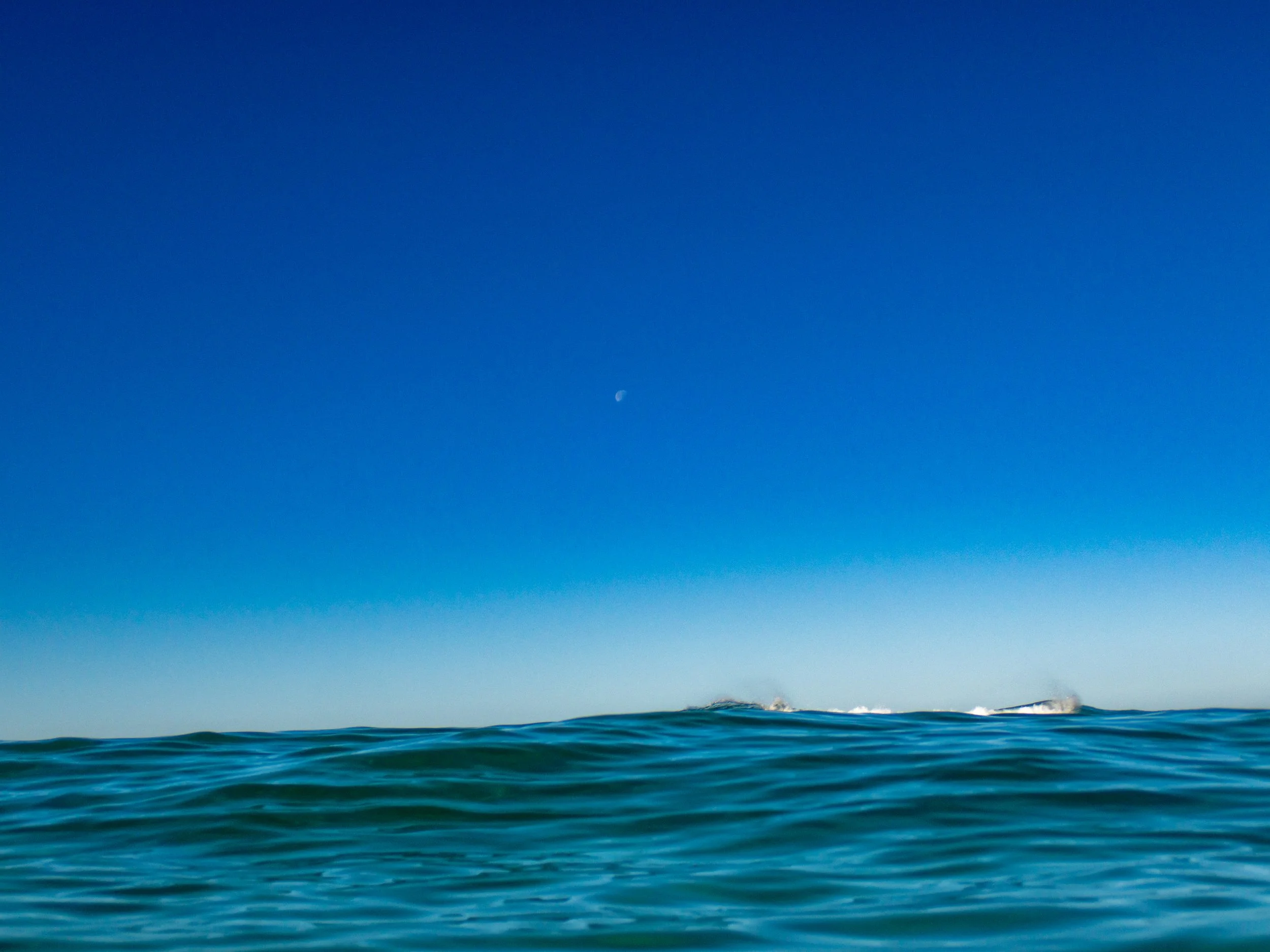 Ocean waves under a clear blue sky with a visible moon in the distance.