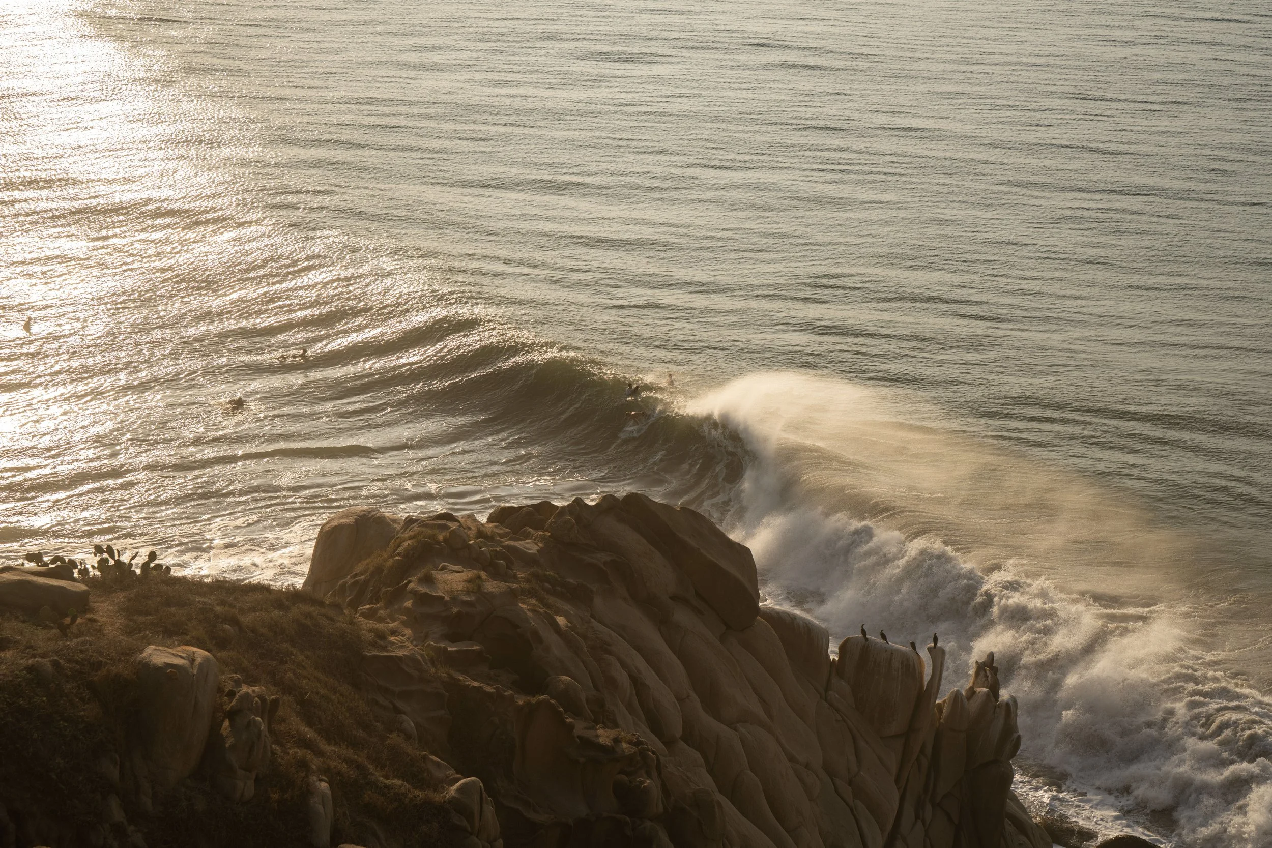 Sunset over the ocean with waves crashing against rocks along the shoreline.