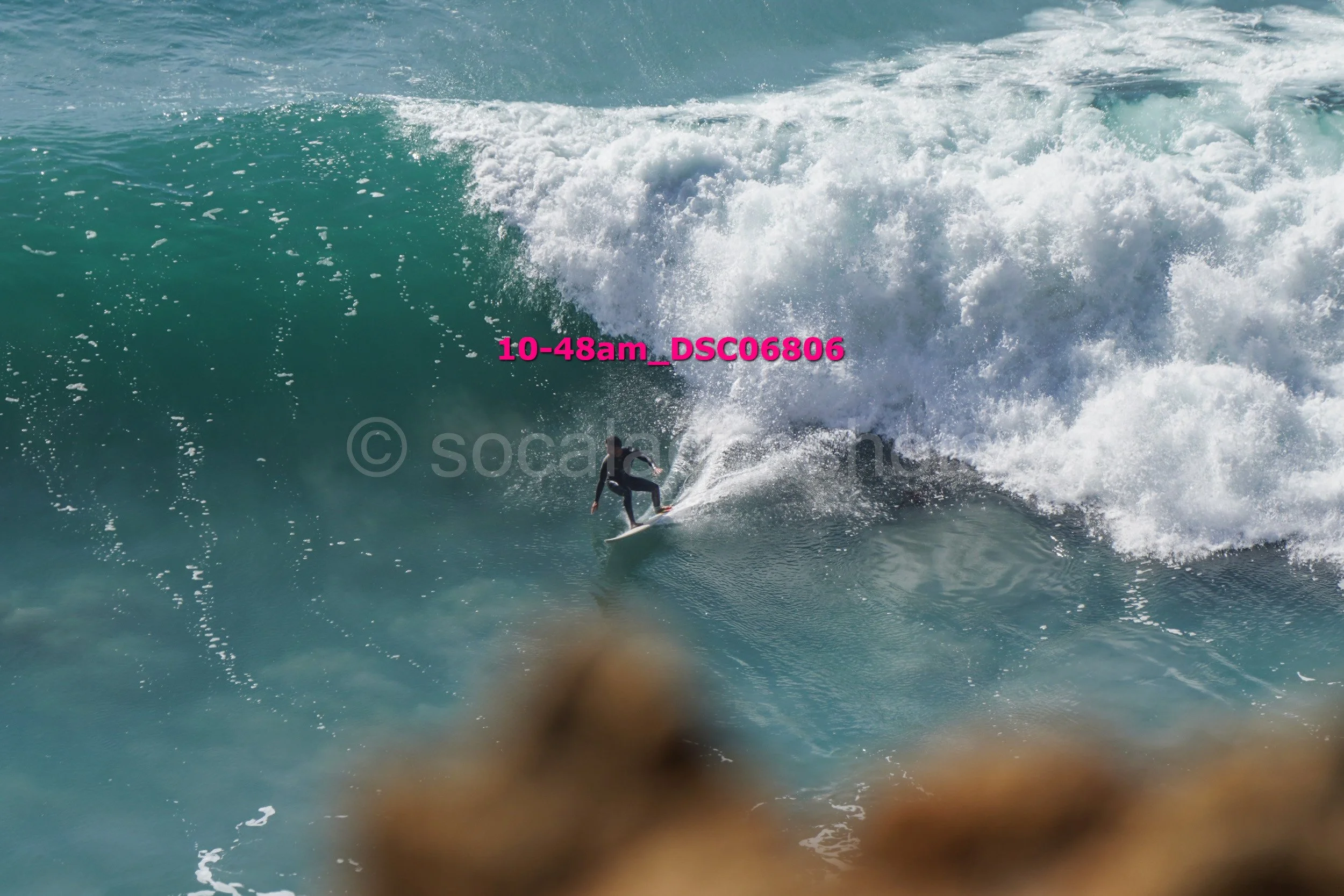 A person surfing on a large ocean wave, with rocks in the foreground and white surf spray around.