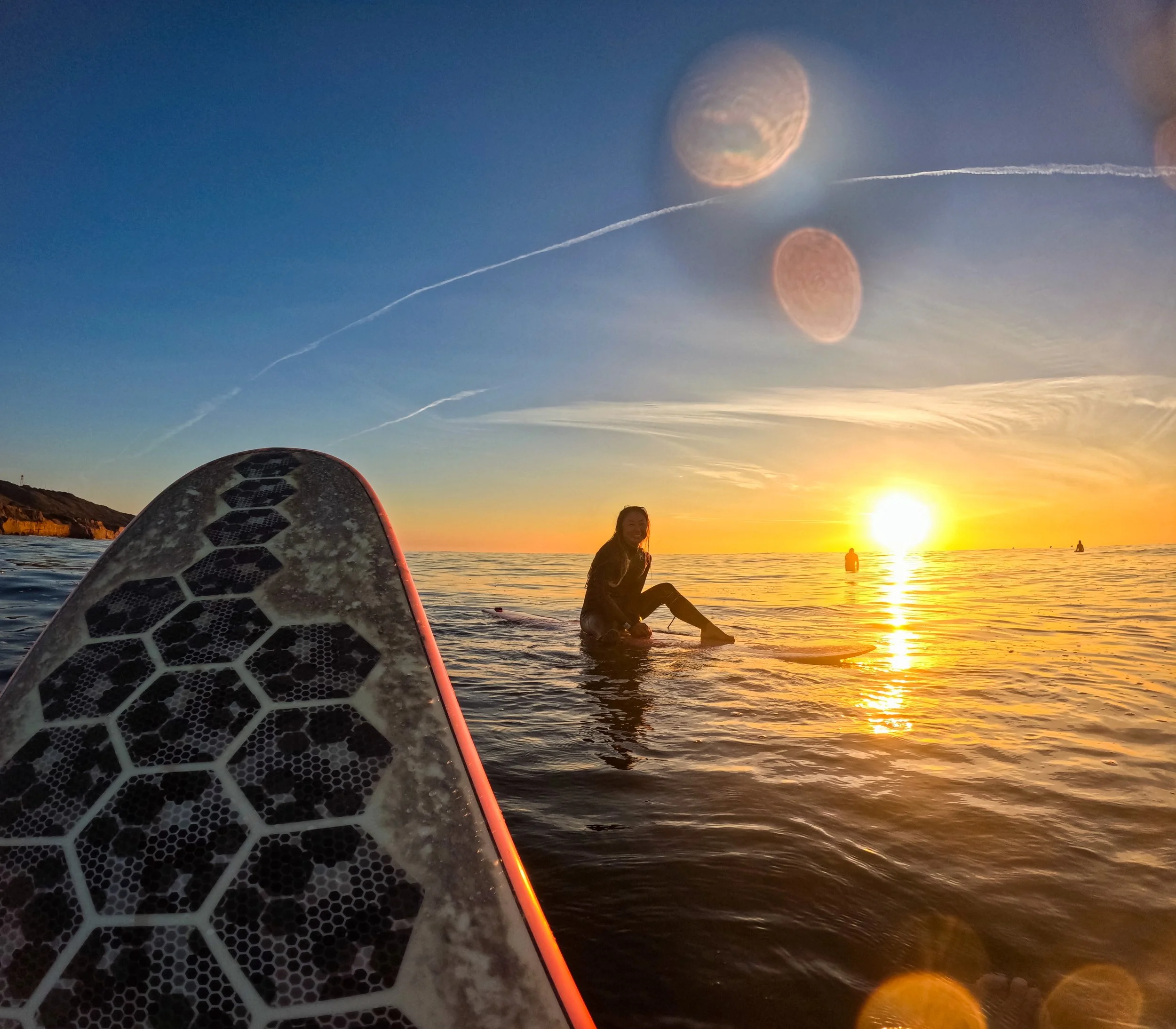 Person sitting on surfboard in ocean during sunset, with other surfers in the distance, and sunlight reflecting on the water.