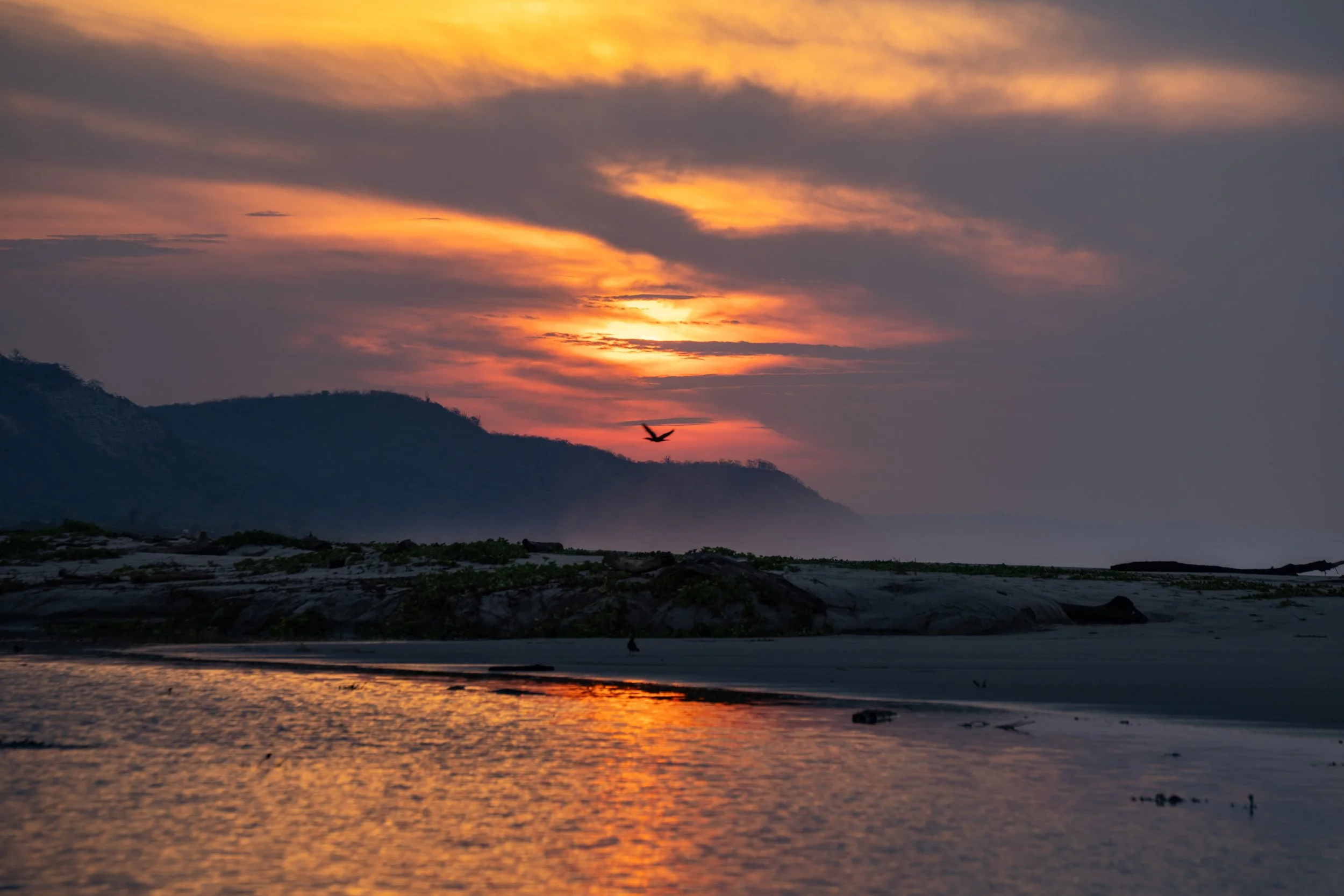 A sunset over a mountain landscape with a bird flying in the sky, reflecting on the water below.
