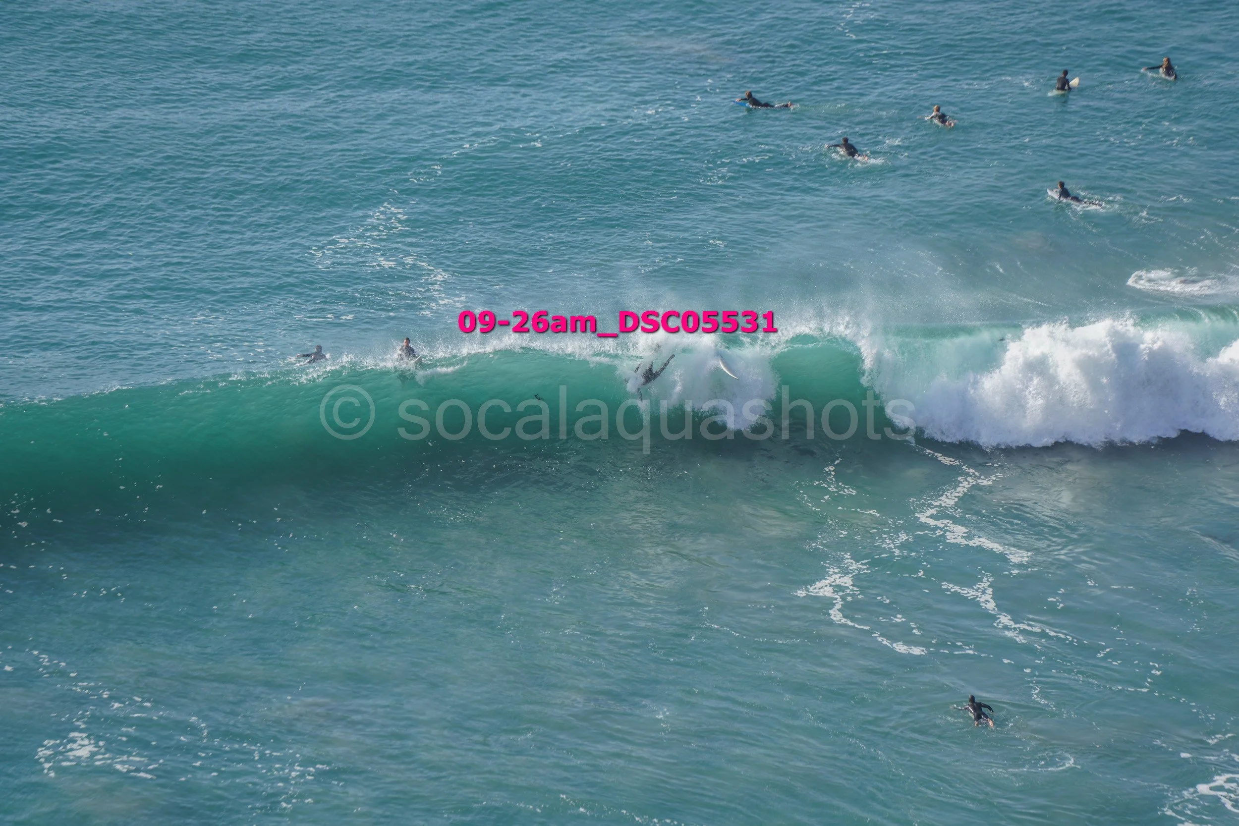 Surfers riding a large wave in the ocean with others waiting or swimming nearby.