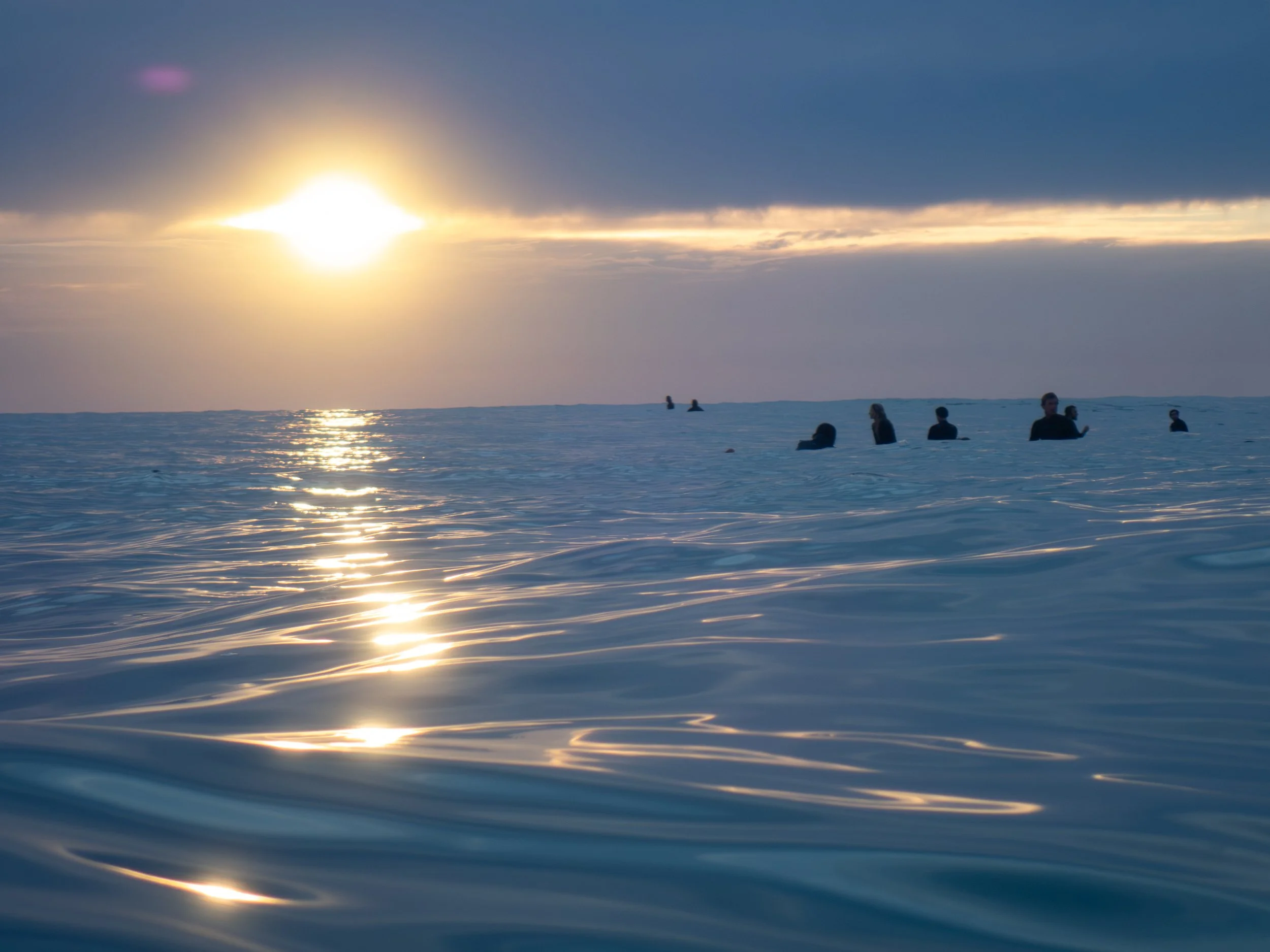 People swimming in the ocean at sunset, with the sun low on the horizon and a partly cloudy sky.