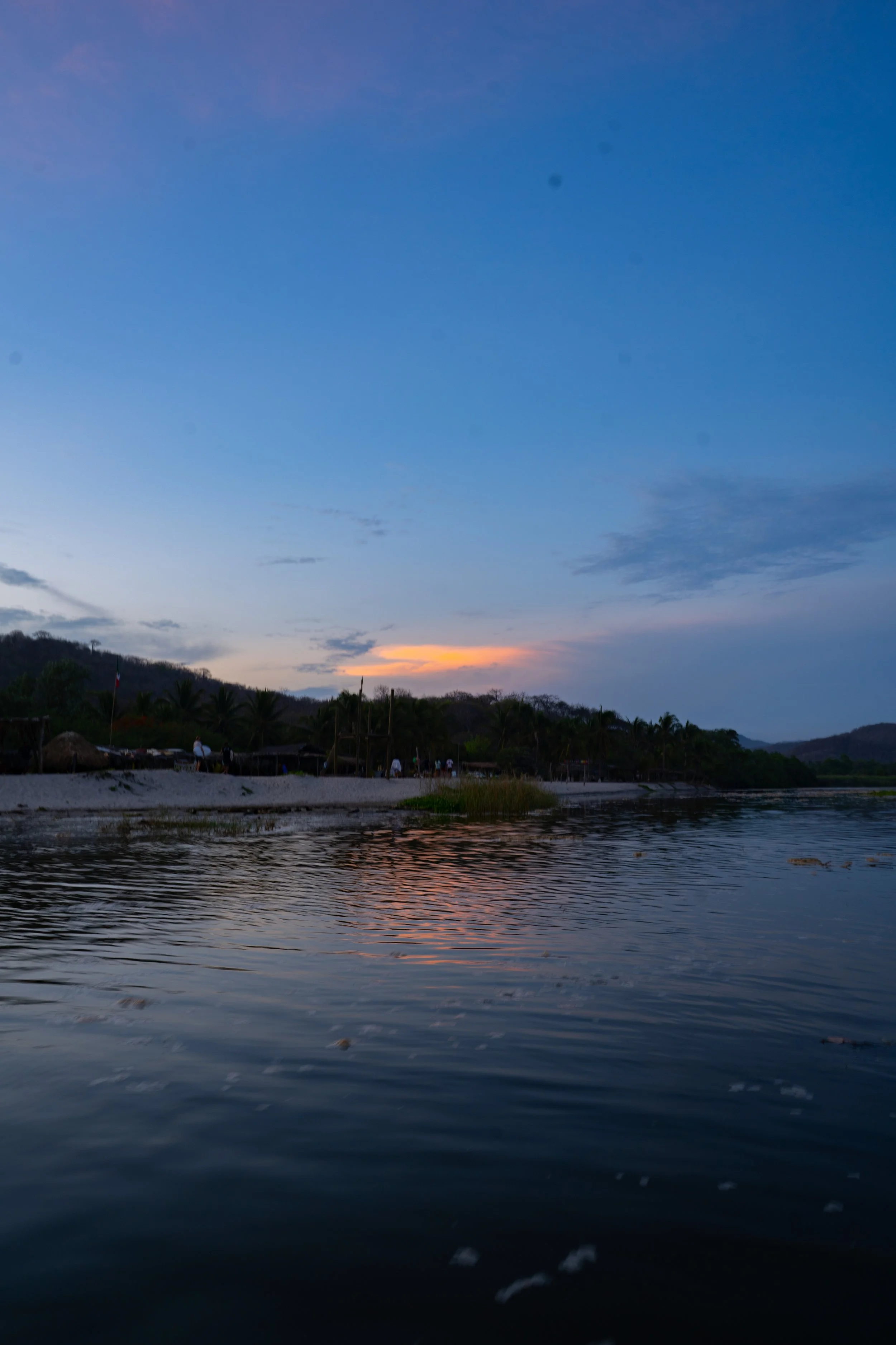 View of a river at sunset with silhouetted trees and a sky transitioning from daylight to dusk.