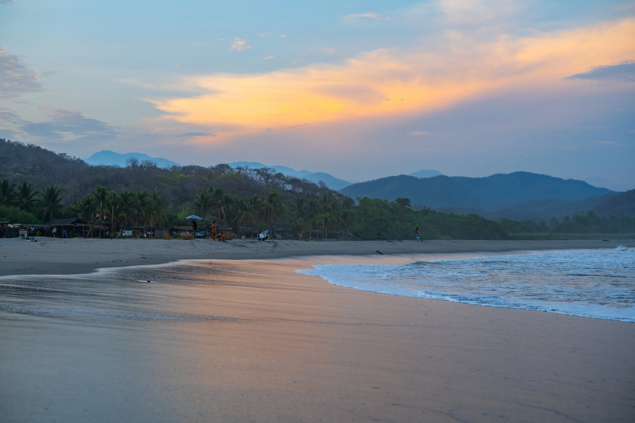 A beach during sunset with calm waves, palm trees, and a few people walking or relaxing near small huts, with mountains in the background and a colorful sky.