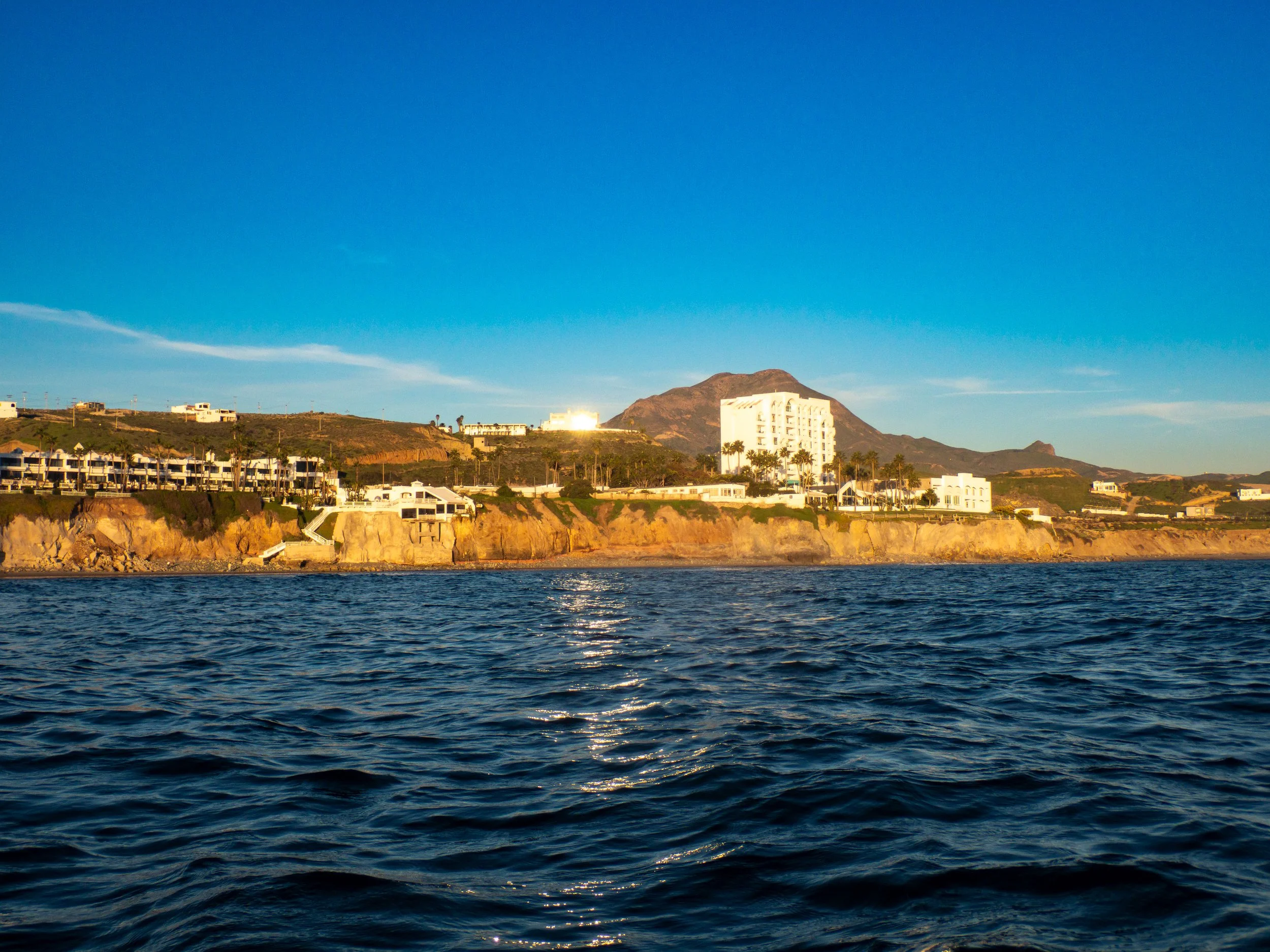 View of a coastal hillside with white buildings, cliffs, and a mountain in the background under a clear blue sky