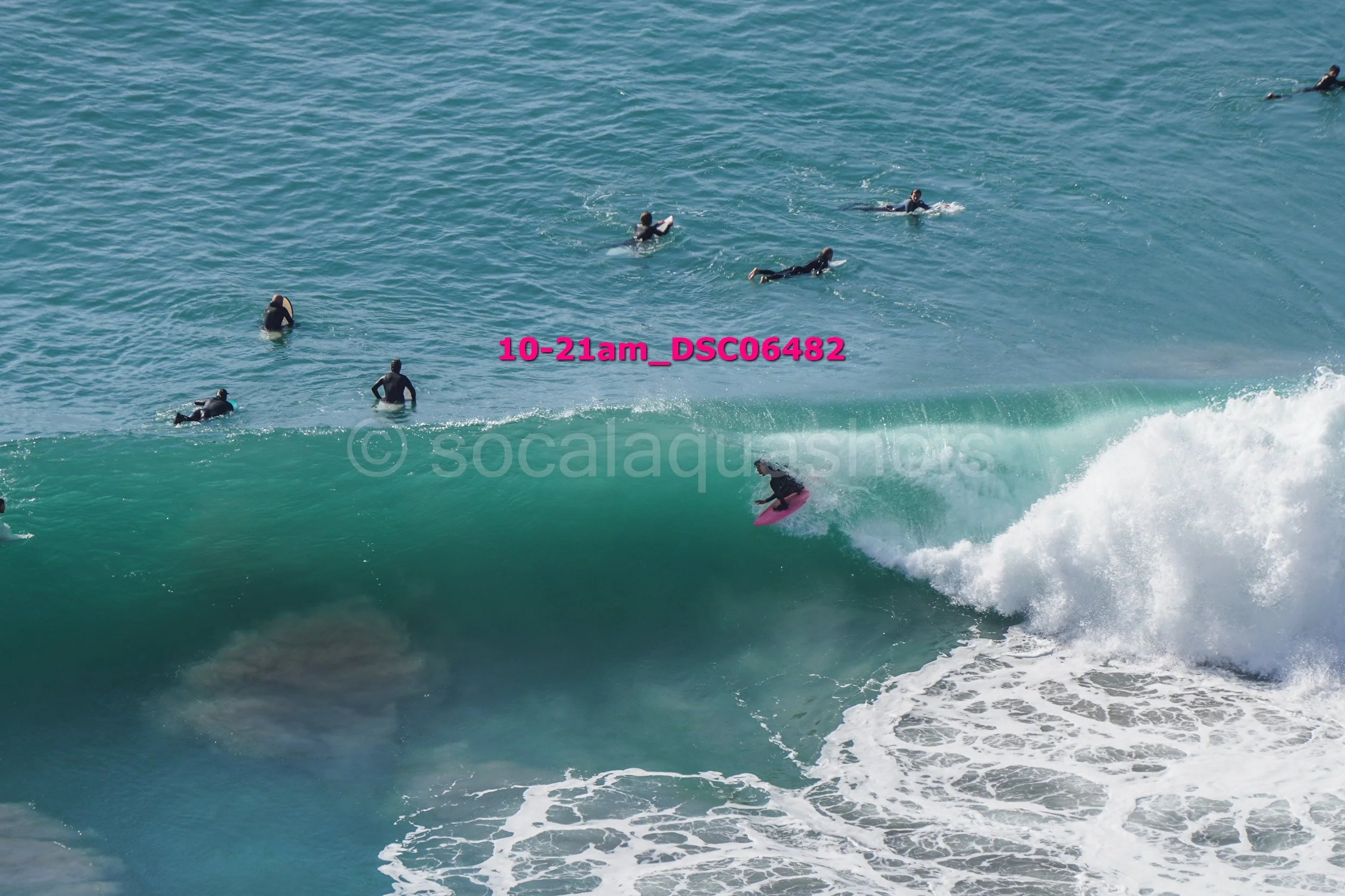 A person surfing on a pink surfboard riding a large wave, with multiple surfers in the water watching nearby.