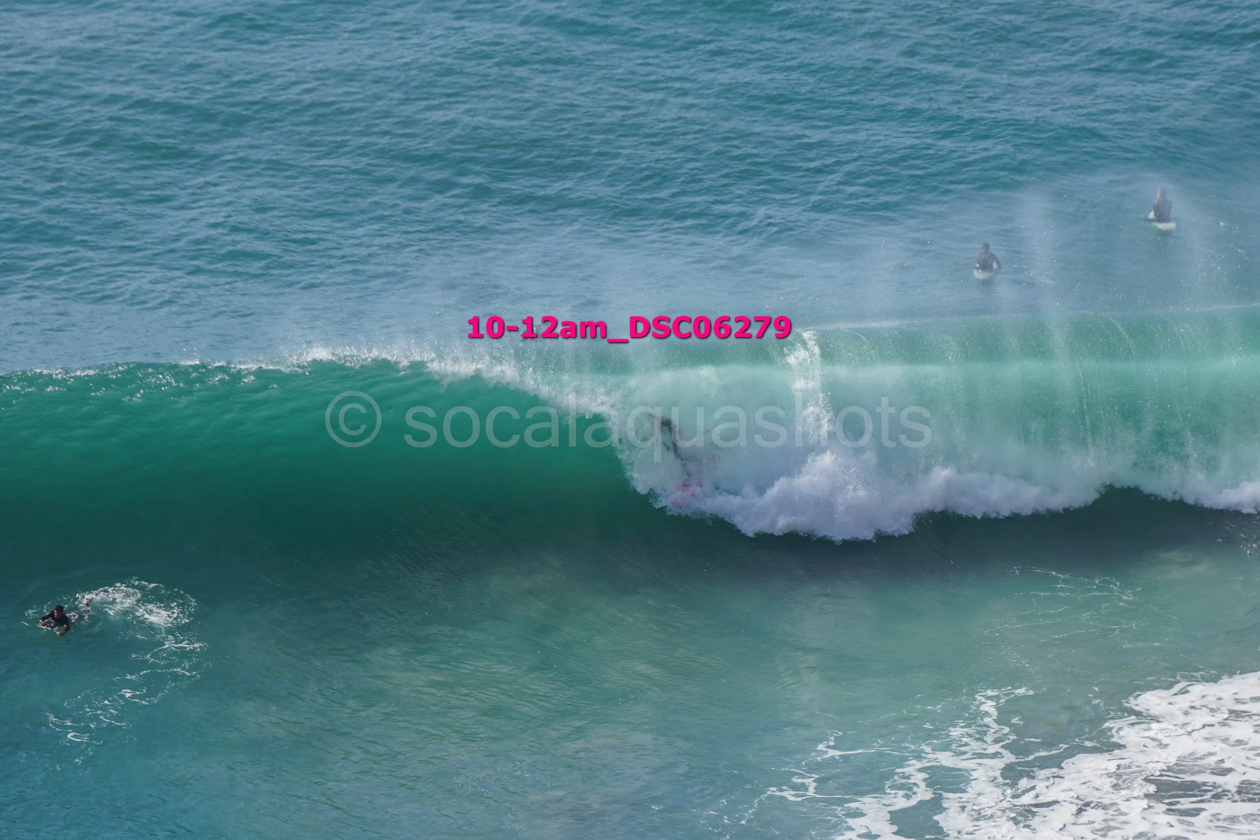 Surfers riding a large wave in the ocean, with some surfers waiting in the water and others on their boards.