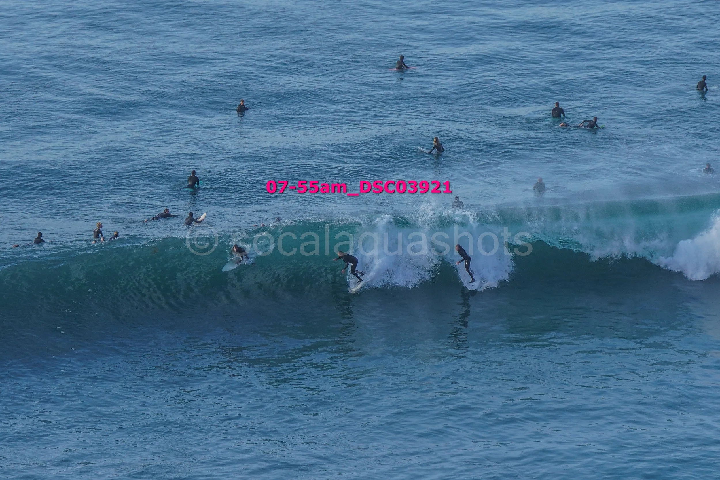 Surfers riding a wave with many people in the water in the background.