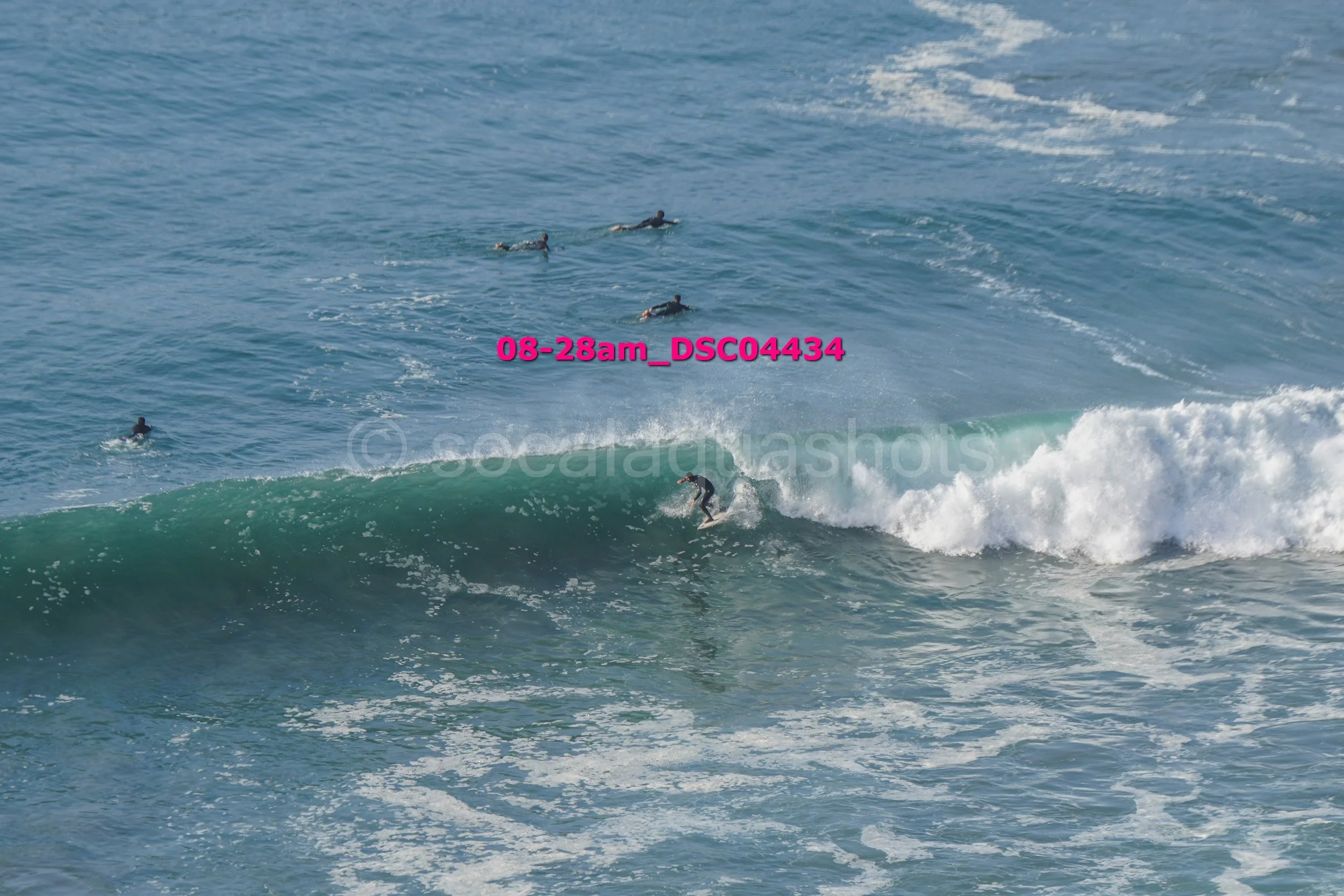 Surfer riding a wave with several people swimming or paddling in the water nearby.