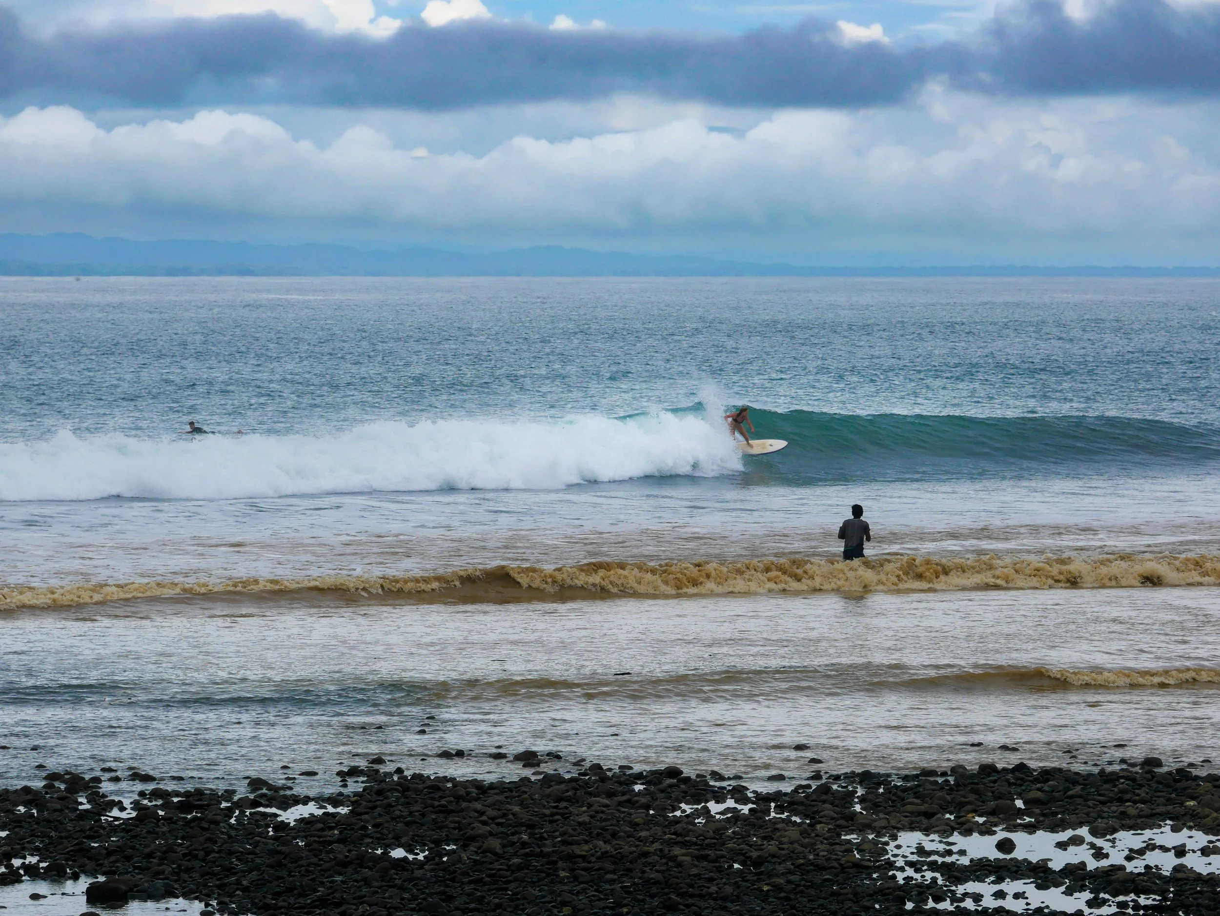 Surfer riding a wave near the shore with a person standing on a rocky beach watching.