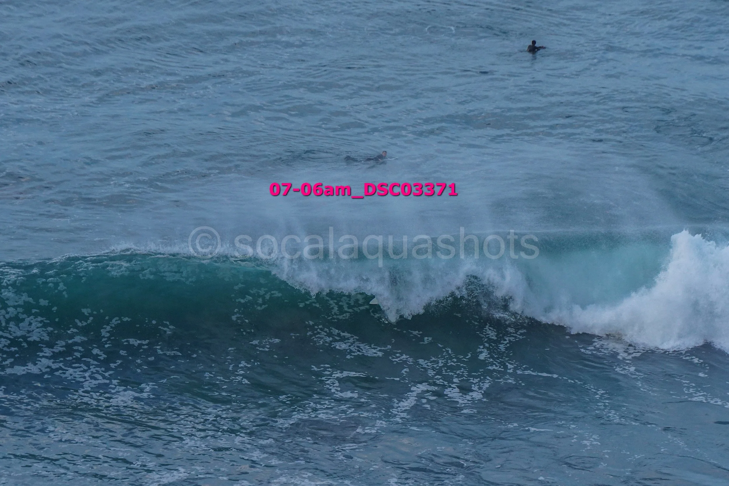 Ocean waves with foam and three surfers in the water in the background.