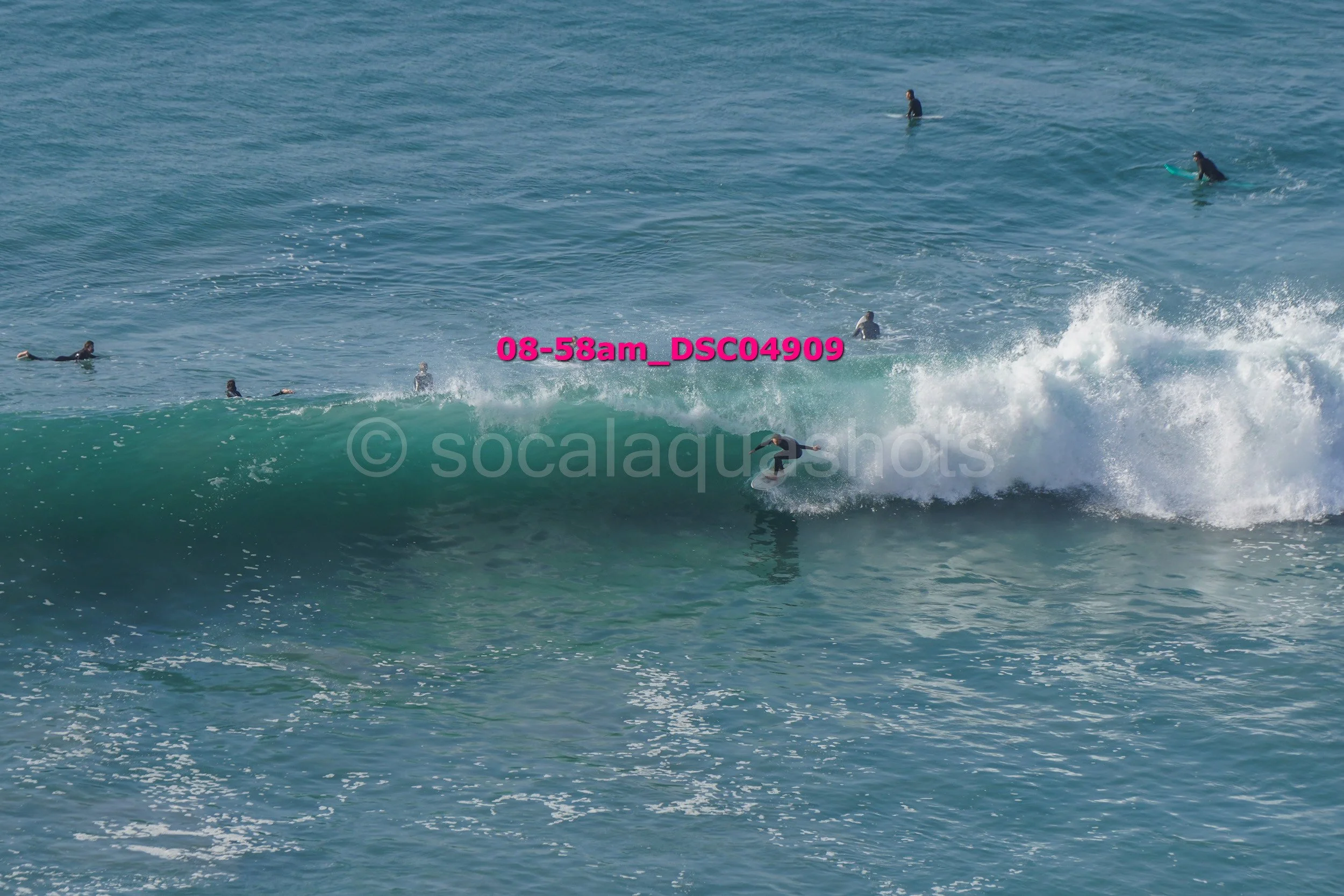 Surfers in the ocean, one riding a wave while others wait or paddle in the water.