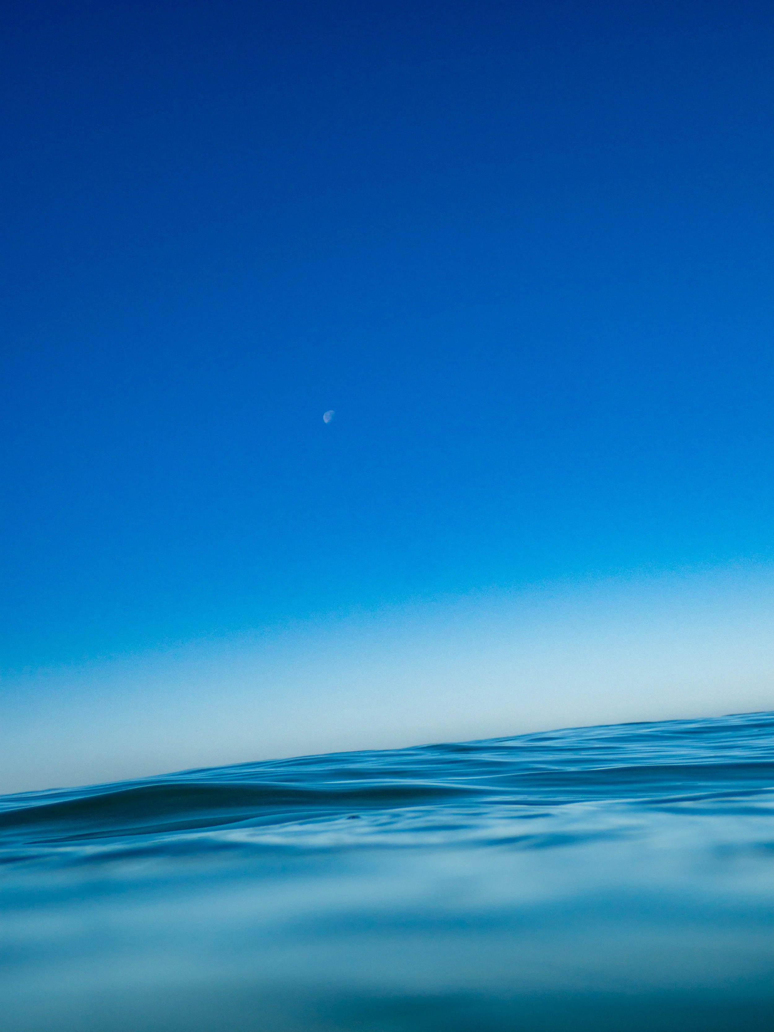 Calm ocean water with a clear blue sky and visible moon.