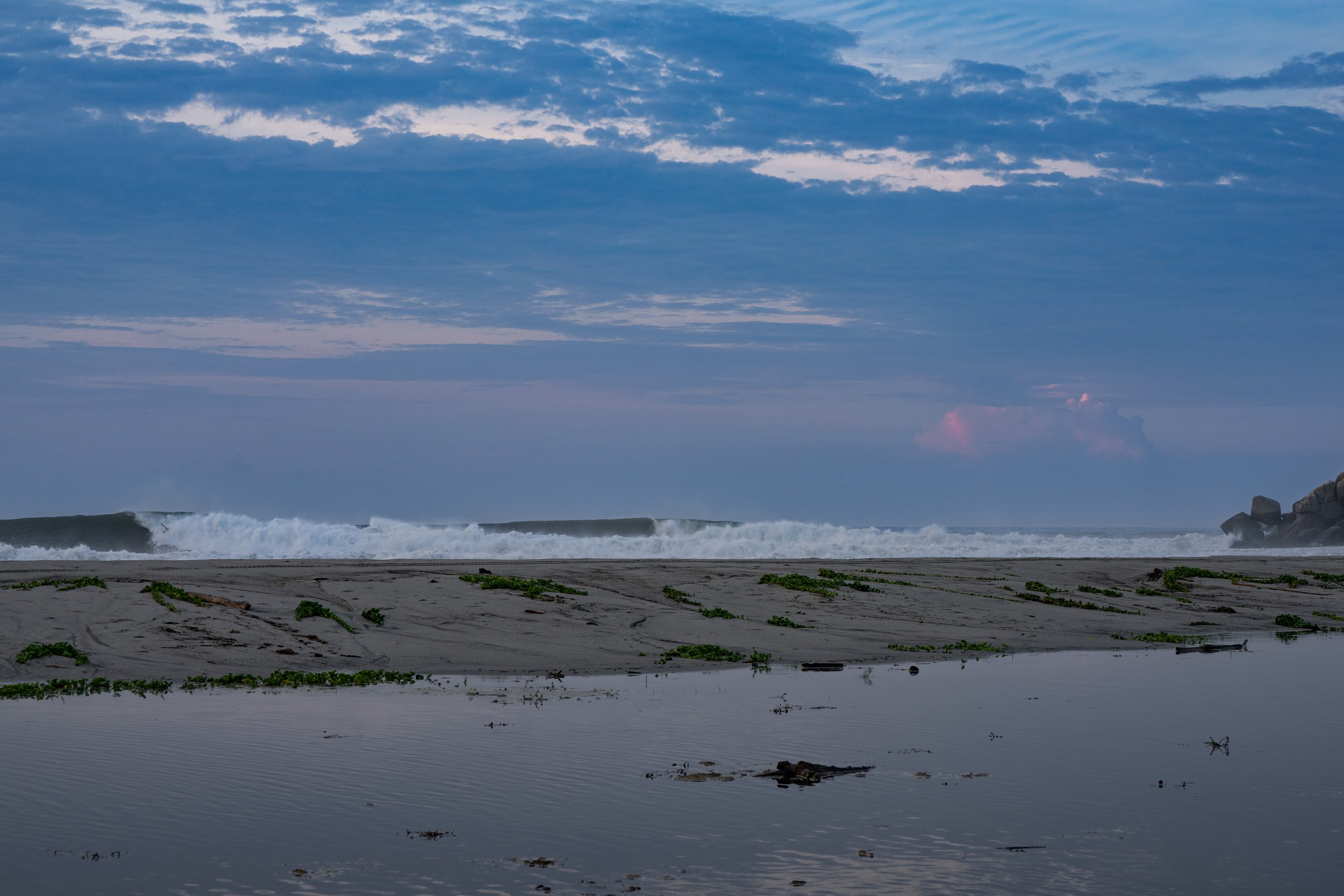 The image shows a beach with waves crashing onto the shore, a cloudy sky above, and distant rocks on the right side.