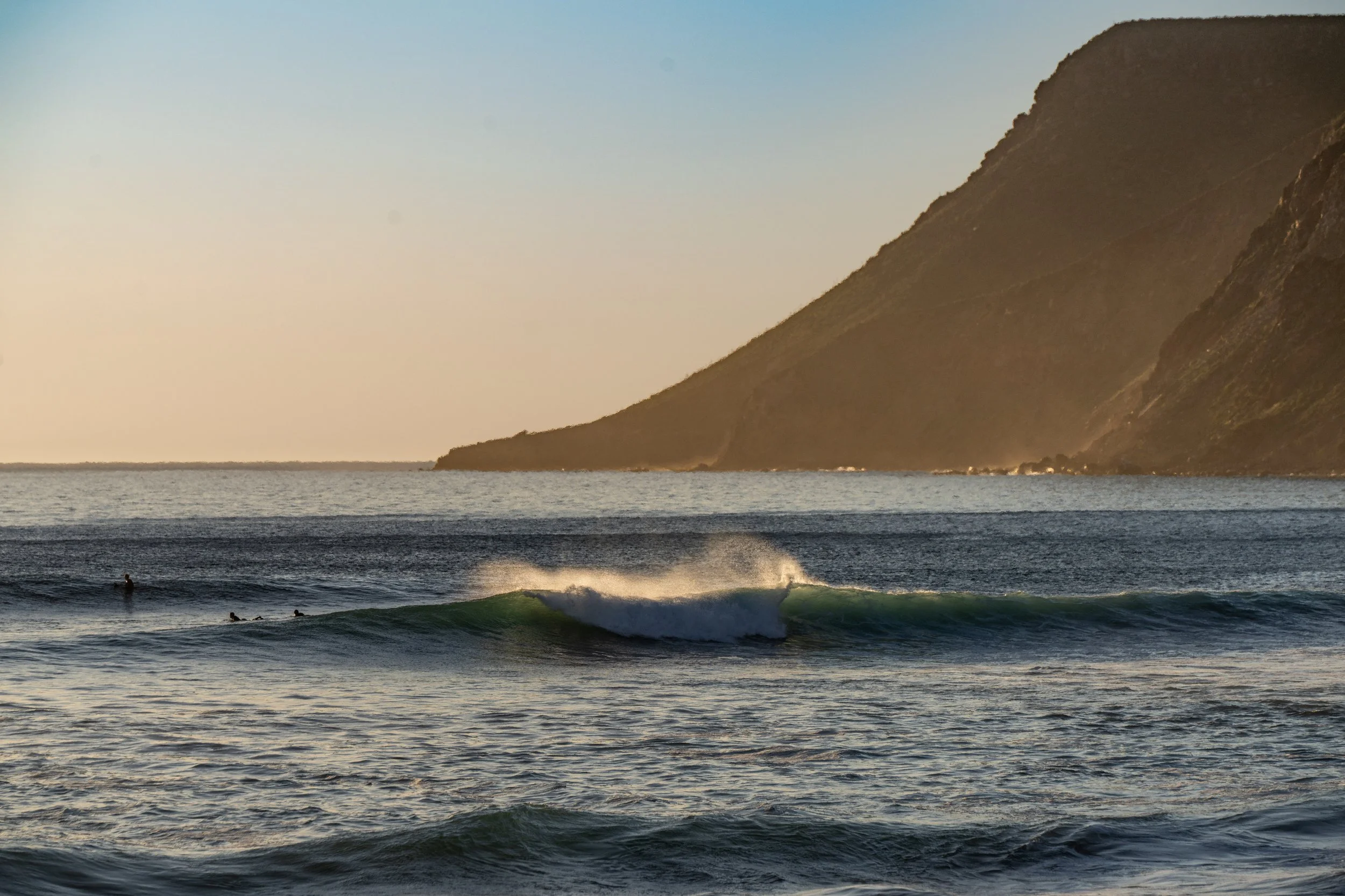 Sea waves with surfers, a mountain on the horizon, and a clear sky at sunset.