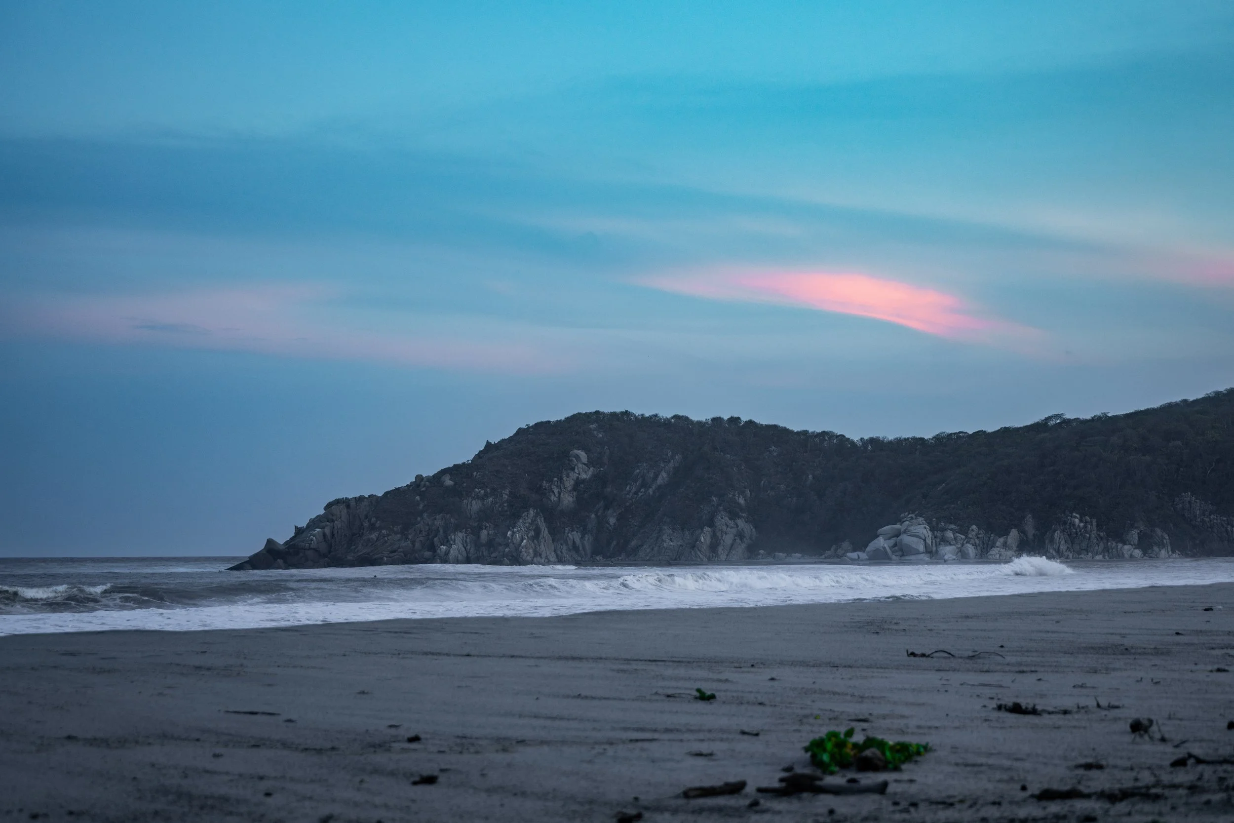 A scenic view of a beach with waves washing onto the shore, a rocky cliff in the background, and a colorful pink and blue sky during sunset or sunrise.