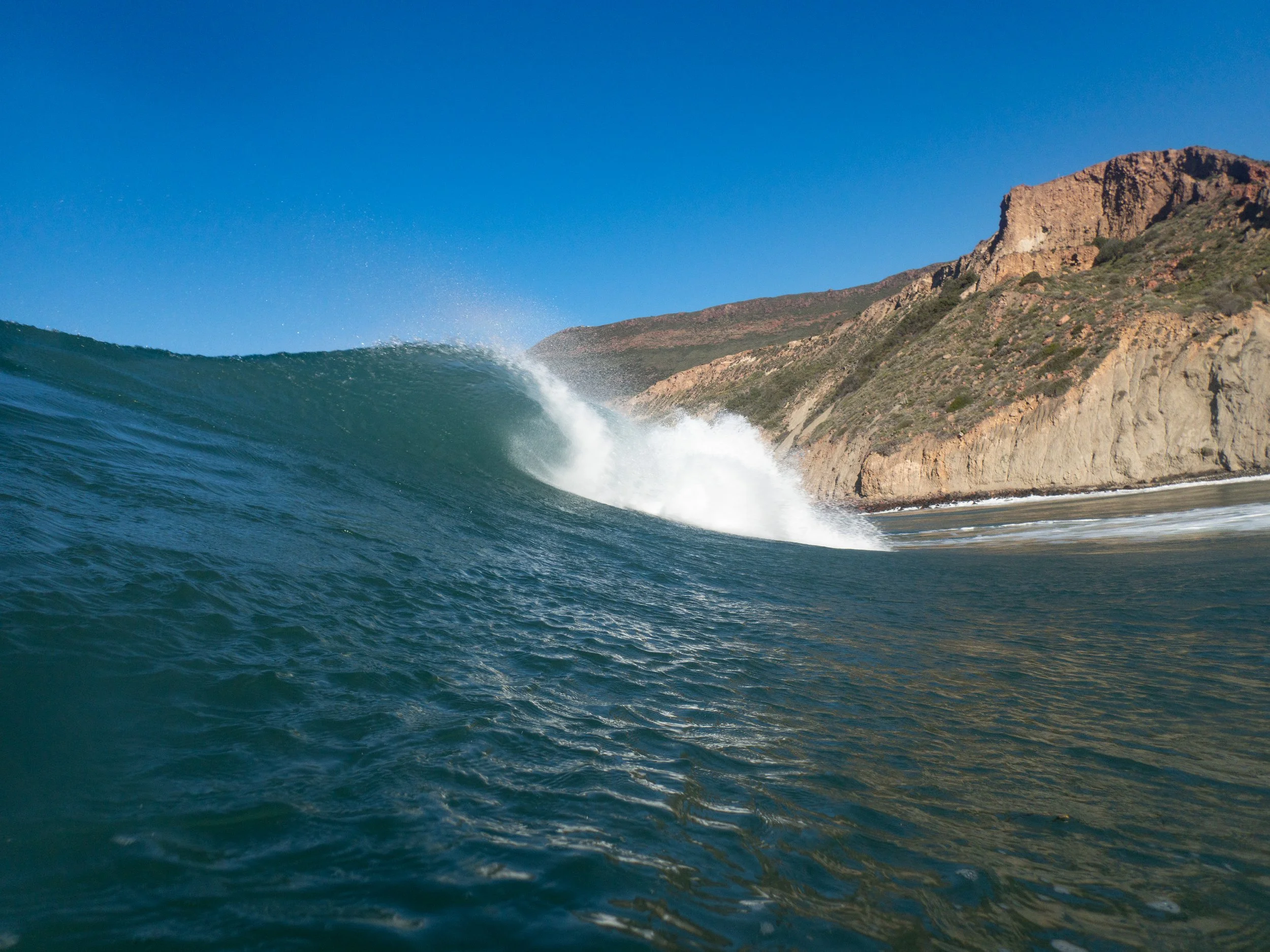 A large ocean wave with a rocky cliff in the background under a clear blue sky.