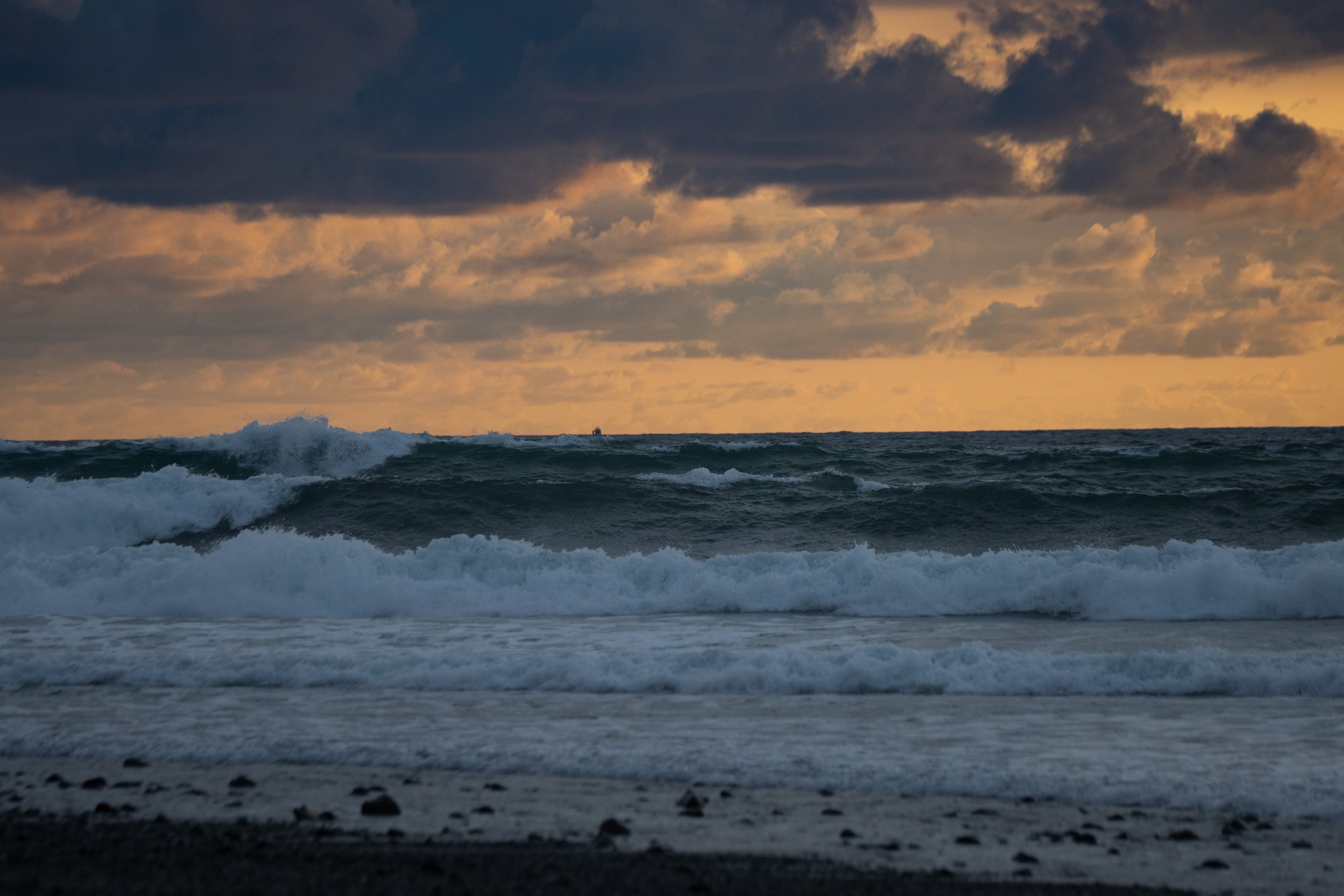 Ocean waves crashing near a sandy beach under a cloudy sky at sunset.
